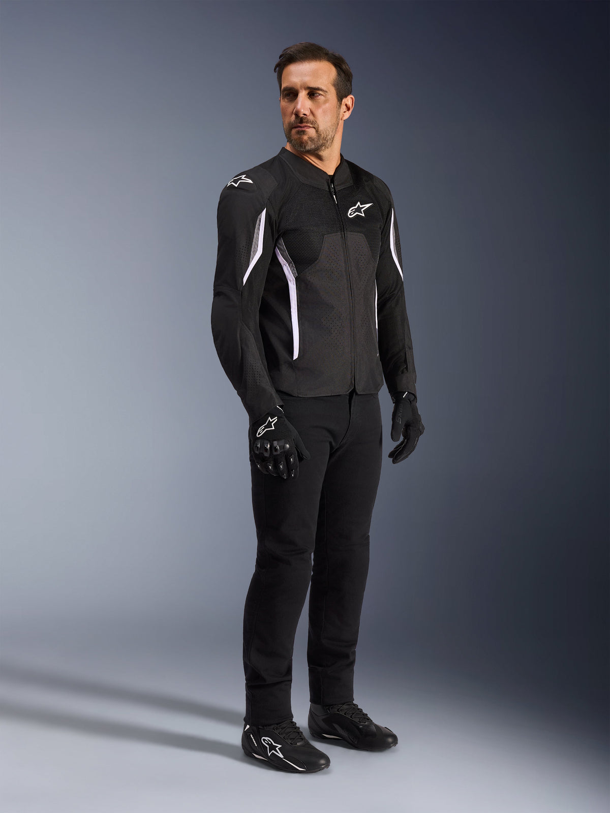 Kurtka Alpinestars AST V3 Air, czarna z białymi akcentami i logotypami, lekka kurtka motocyklowa z wentylacją siatkową, noszona przez mężczyznę z czarnymi spodniami, rękawicami i butami motocyklowymi, techniczna letnia odzież do jazdy sportowej i miejskiej.