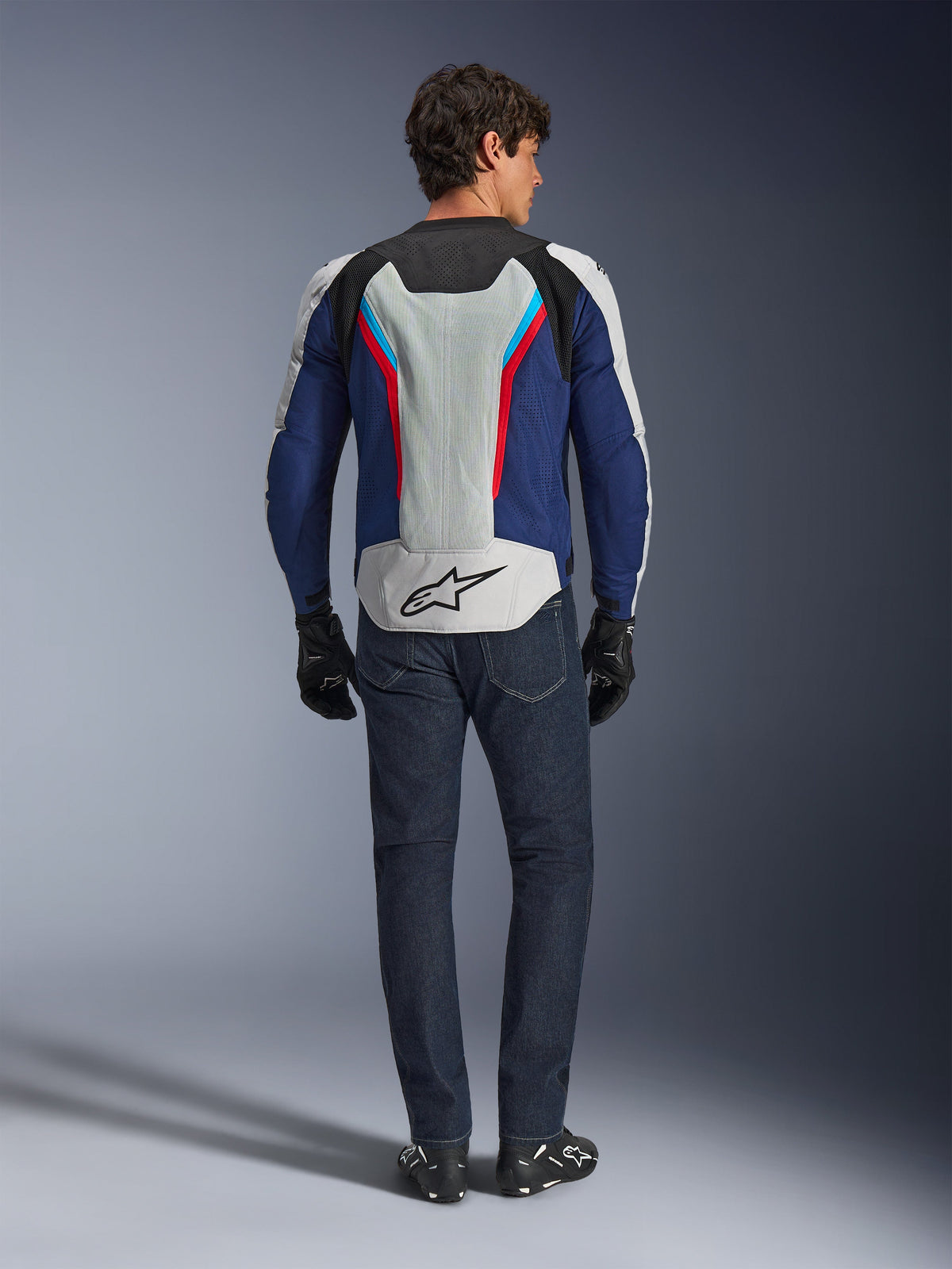 Kurtka Alpinestars AST V3 Air, widok z tyłu, w kolorystyce srebrnej, granatowej i jaskrawoczerwonej, z dużym panelem wentylacyjnym z siatki na plecach i logo Alpinestars, noszona z ciemnoniebieskimi dżinsami i czarnymi rękawicami, sportowa odzież motocyklowa na ciepłe dni
