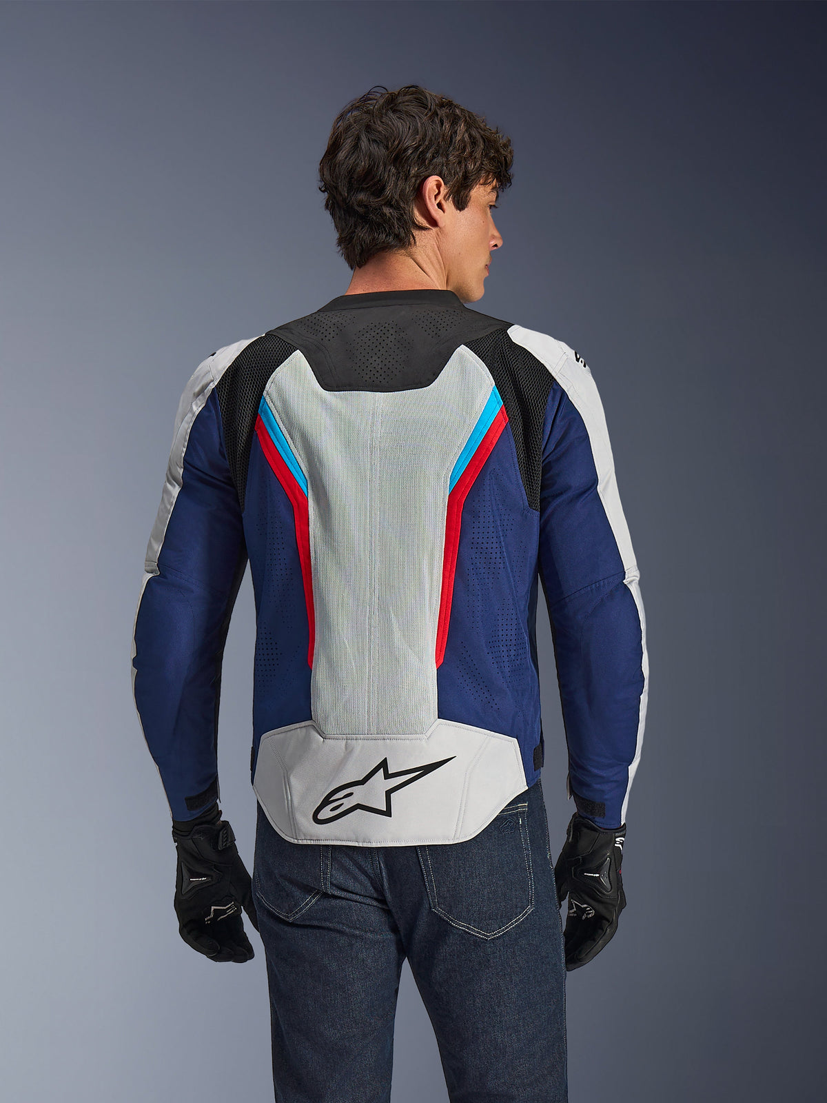 Kurtka Alpinestars AST V3 Air, kurtka motocyklowa, srebrno-granatowo-czerwona, widok z tyłu na motocykliście, wyposażona w panele z siatki technicznej i laserowe perforacje dla lepszej wentylacji, czarne logo gwiazdy w dolnej części pleców, sportowy design na ciepłe dni