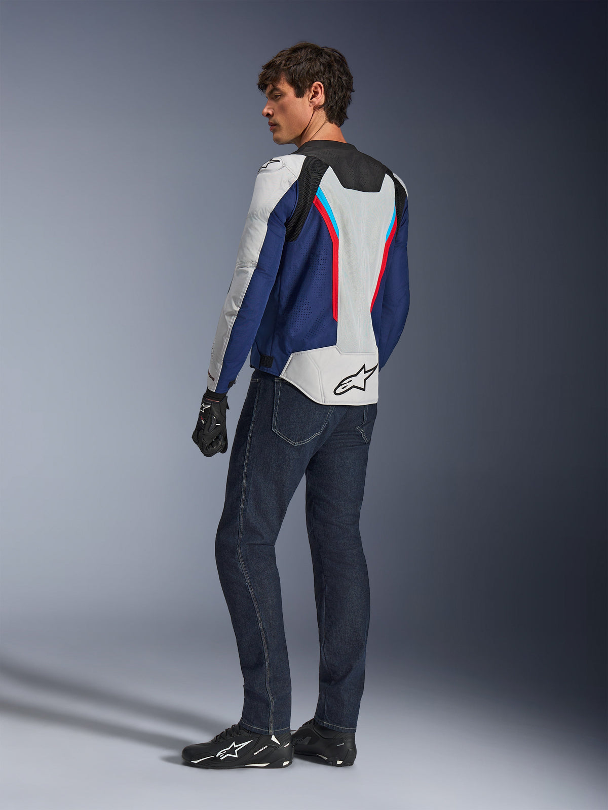 Alpinestars AST V3 Air Jacket, kurtka motocyklowa w kolorze srebrnym, granatowym i jasnoczerwonym, widok z tyłu, zestawiona z ciemnoniebieskimi dżinsami motocyklowymi i czarnym ekwipunkiem, oddychające panele z siatki, logo Alpinestars na plecach, techniczna odzież o sportowym kroju