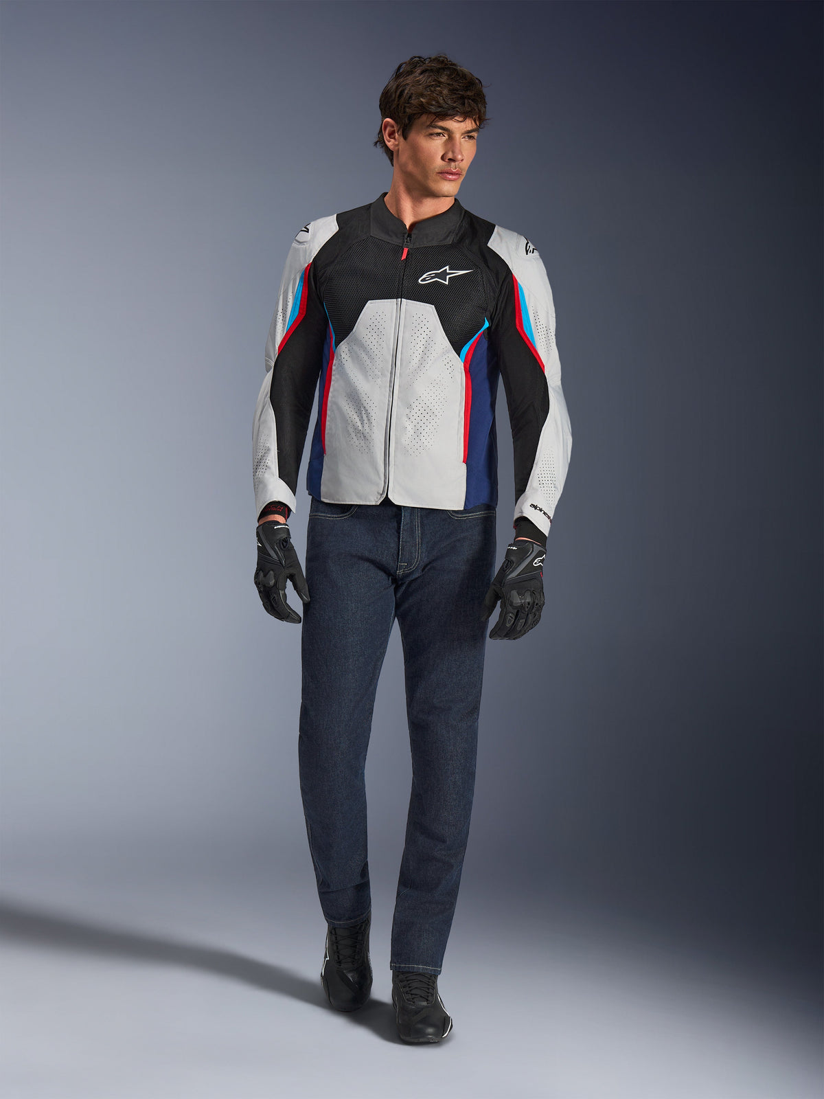 Męski model w kurtce Alpinestars AST V3 Air w kolorze Silver Deep Blue Bright Red, wyposażonej w wentylację siatkową i perforowane panele, w połączeniu z ciemnymi dżinsami motocyklowymi i czarnymi rękawicami; sportowa odzież motocyklowa na ciepłe dni.