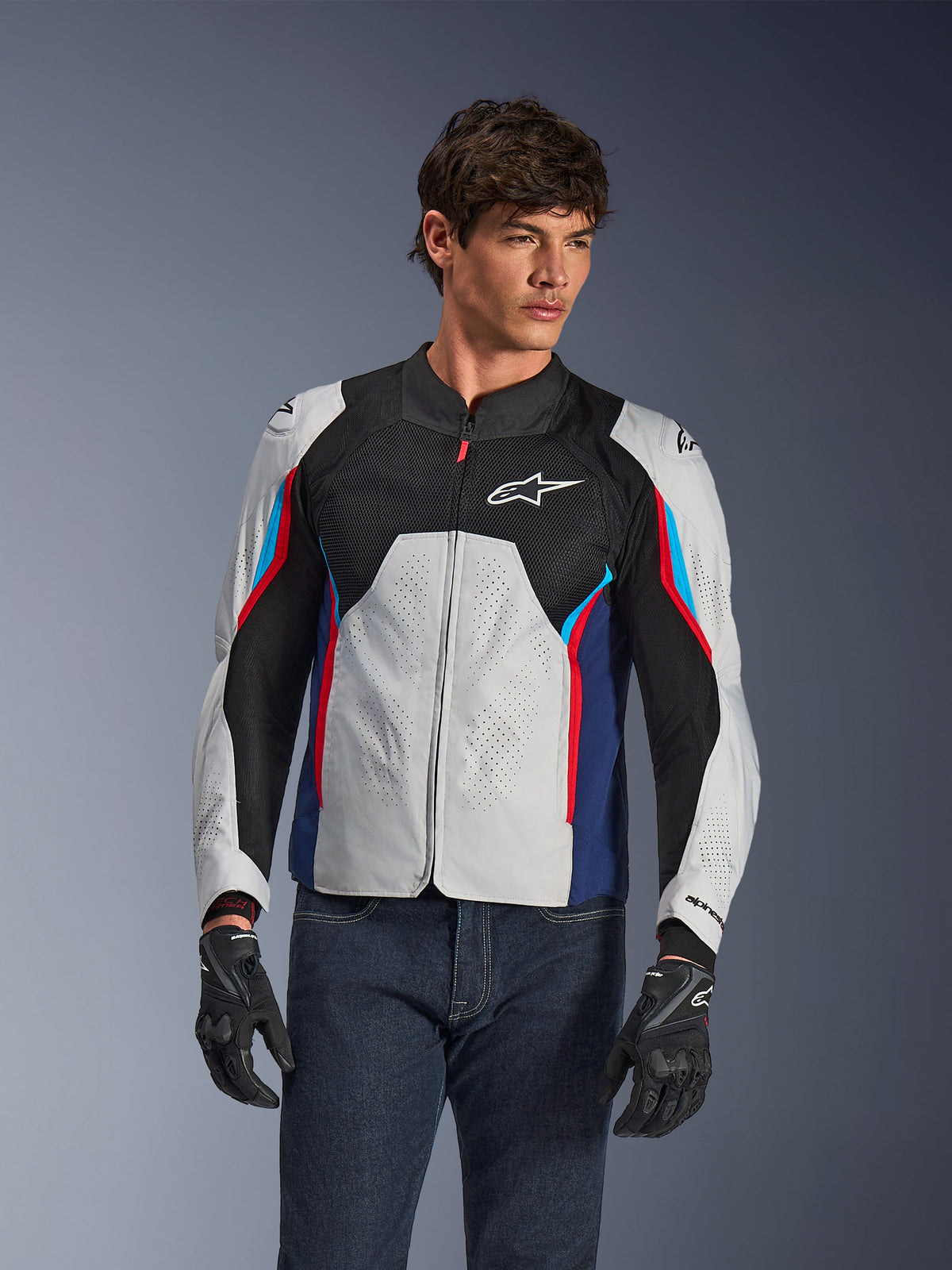 Kurtka Alpinestars AST V3 Air w kolorze srebrnym, granatowym i jasnoczerwonym, noszona przez modela w ciemnych dżinsach i czarnych rękawicach, z perforowanymi srebrnymi panelami, czarną siatką wentylacyjną oraz sportowymi czerwonymi i niebieskimi akcentami.