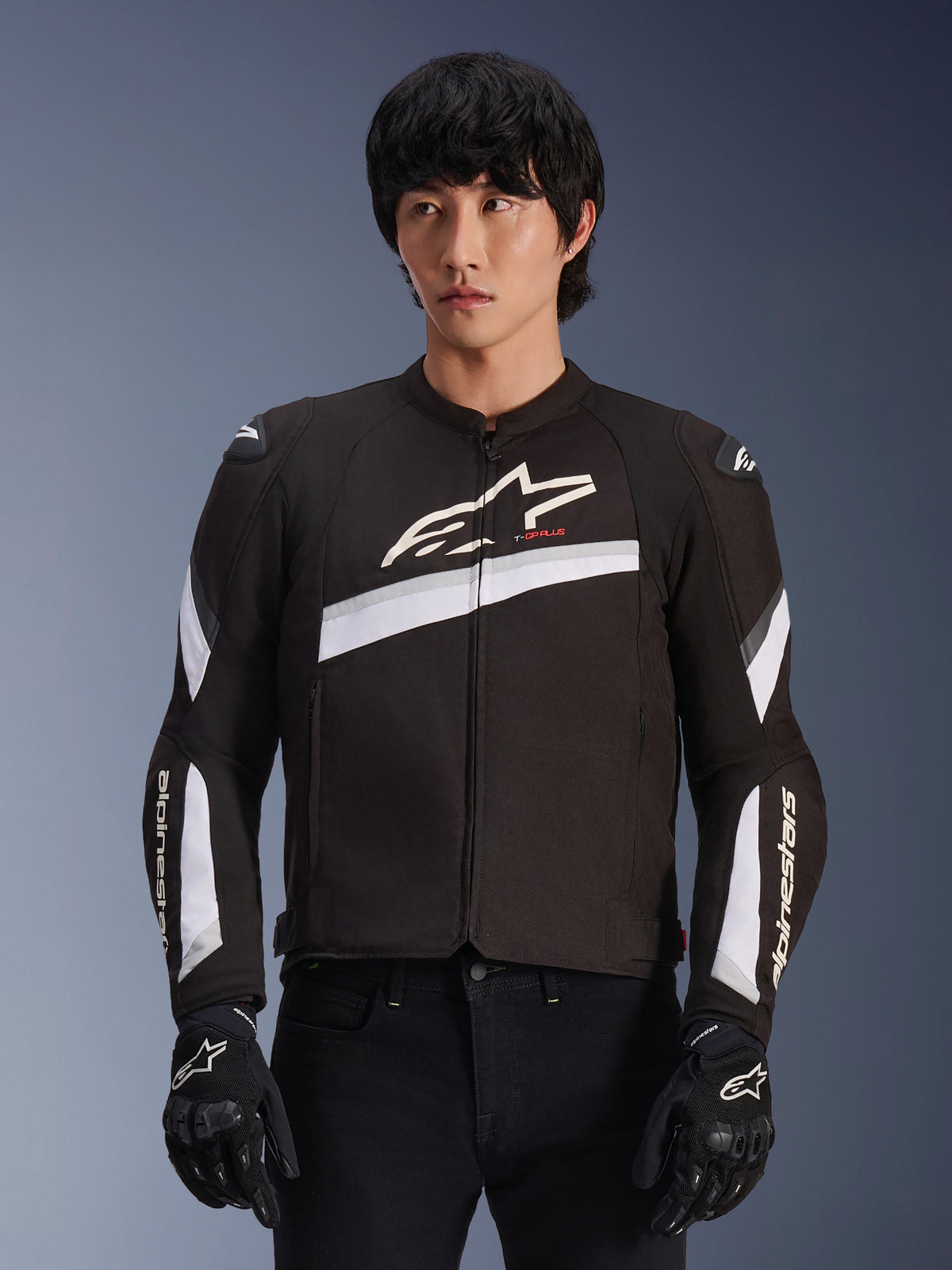 Alpinestars T-GP Plus R V4 Jacket, czarno-biała tekstylna kurtka motocyklowa, białe ukośne akcenty i logo marki na klatce piersiowej oraz rękawach, noszona przez modela w czarnych rękawicach motocyklowych, dopasowany sportowy krój do jazdy szosowej