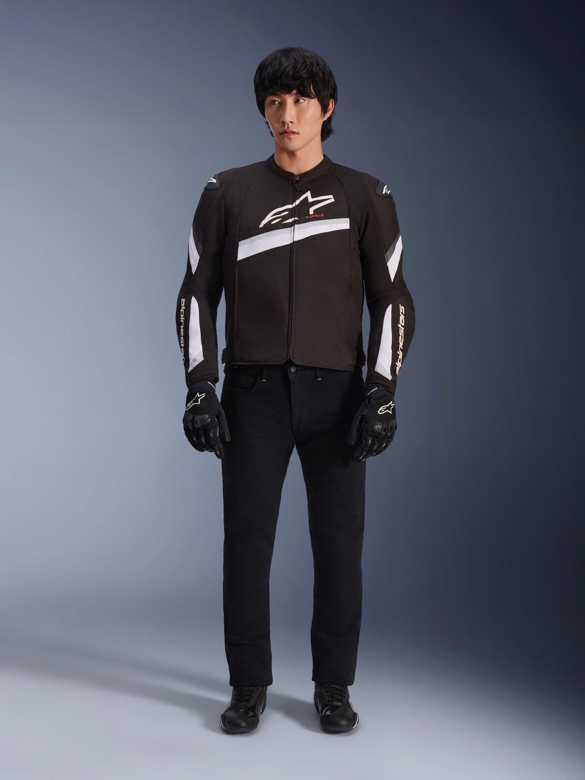 Kurtka Alpinestars T-GP Plus R V4, czarno-biała, sportowa tekstylna kurtka motocyklowa, prezentowana przez modela w czarnych spodniach i rękawicach, z wyraźnym białym logo Alpinestars na klatce piersiowej i rękawach, smukły design inspirowany wyścigami