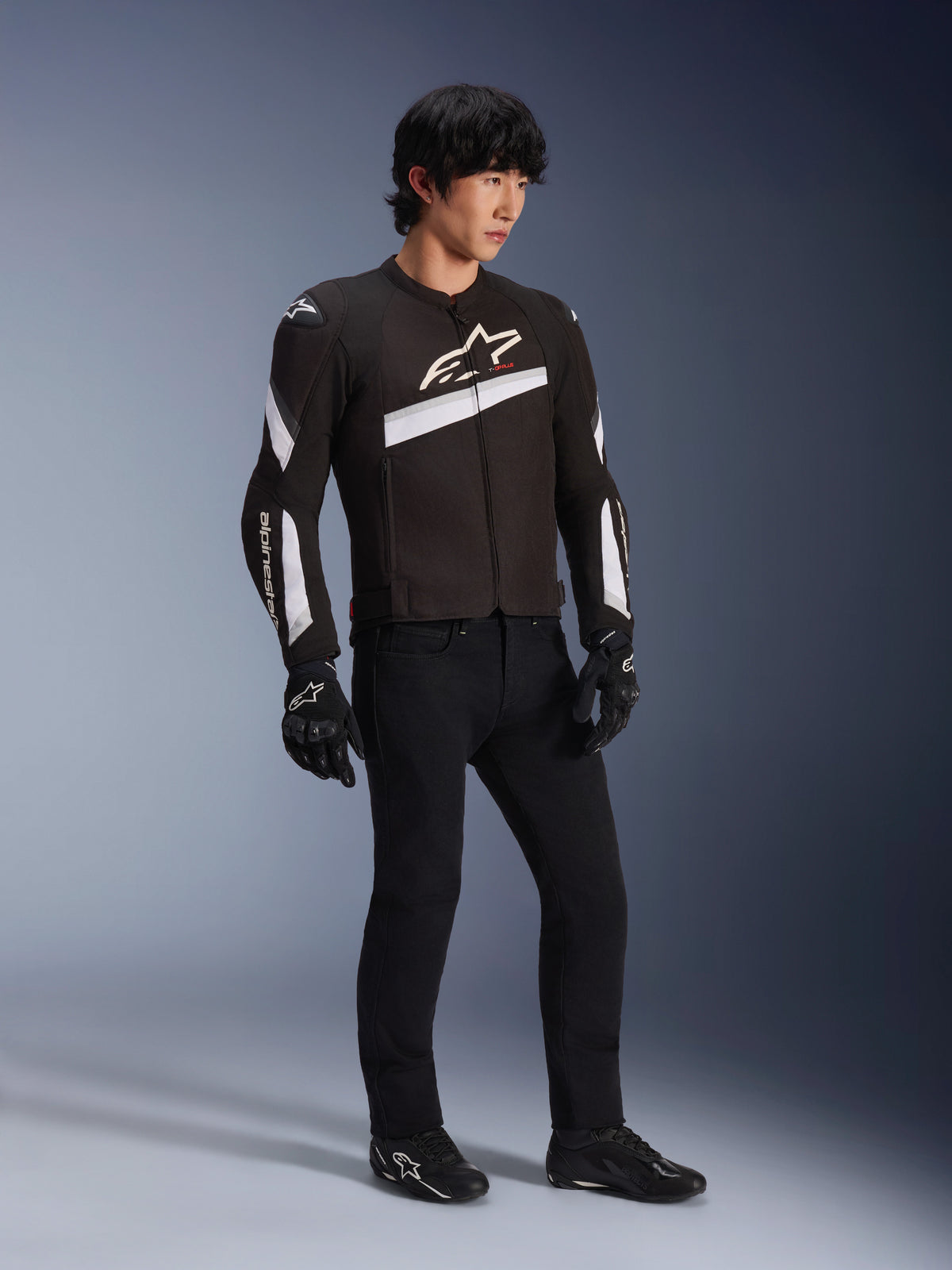 Alpinestars T-GP Plus R V4 Jacket, czarno-biała tekstylna kurtka motocyklowa noszona przez modela z czarnymi spodniami i rękawicami, białe logo Alpinestars na klatce piersiowej i rękawach, sportowy design do jazdy szosowej