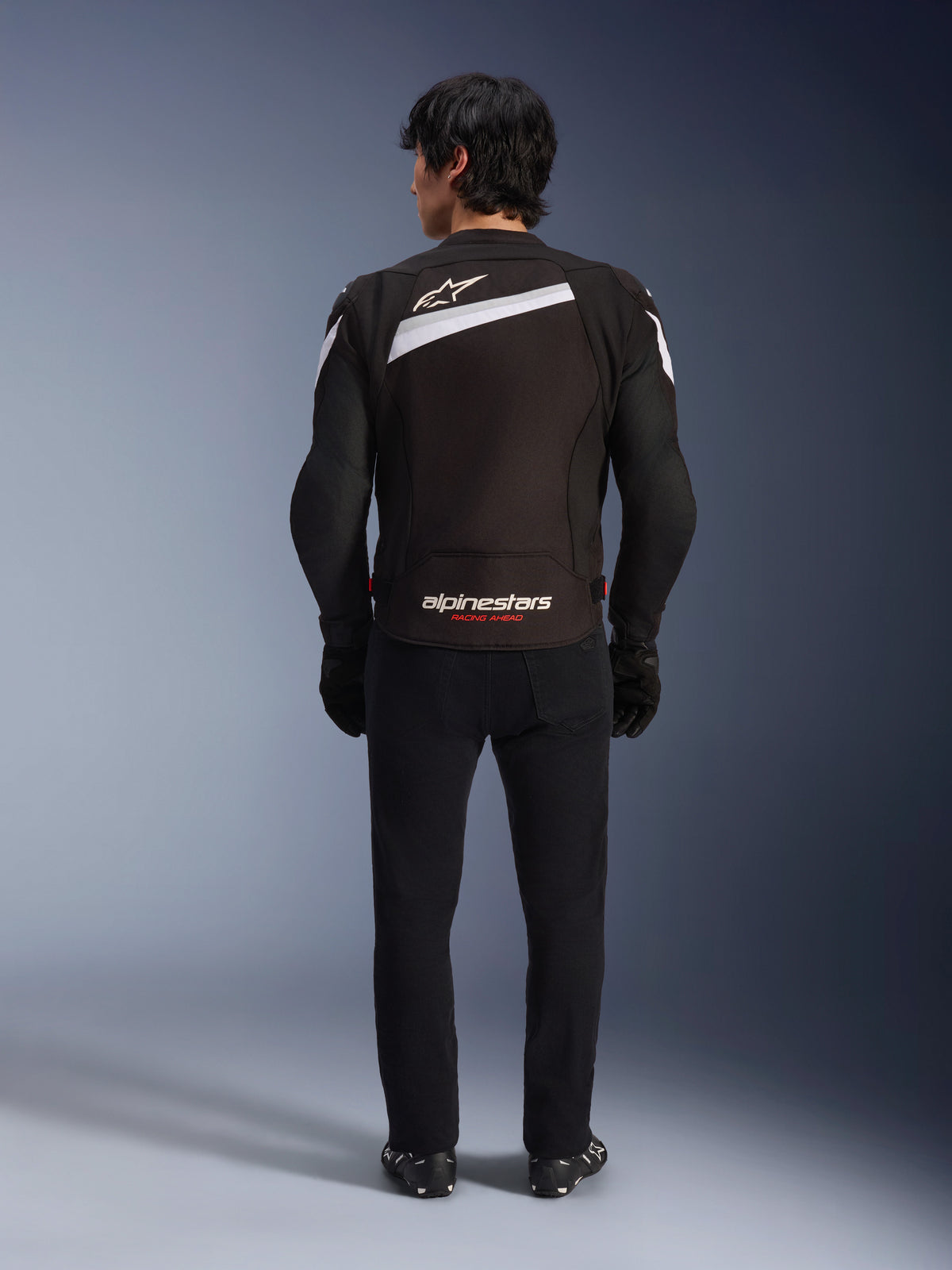 Kurtka Alpinestars T-GP Plus R V4, czarno-biała sportowa tekstylna kurtka motocyklowa, widok z tyłu z białym logo Alpinestars i brandingiem „RACING AHEAD”, noszona z czarnymi spodniami i butami, opływowa konstrukcja do jazdy wyczynowej