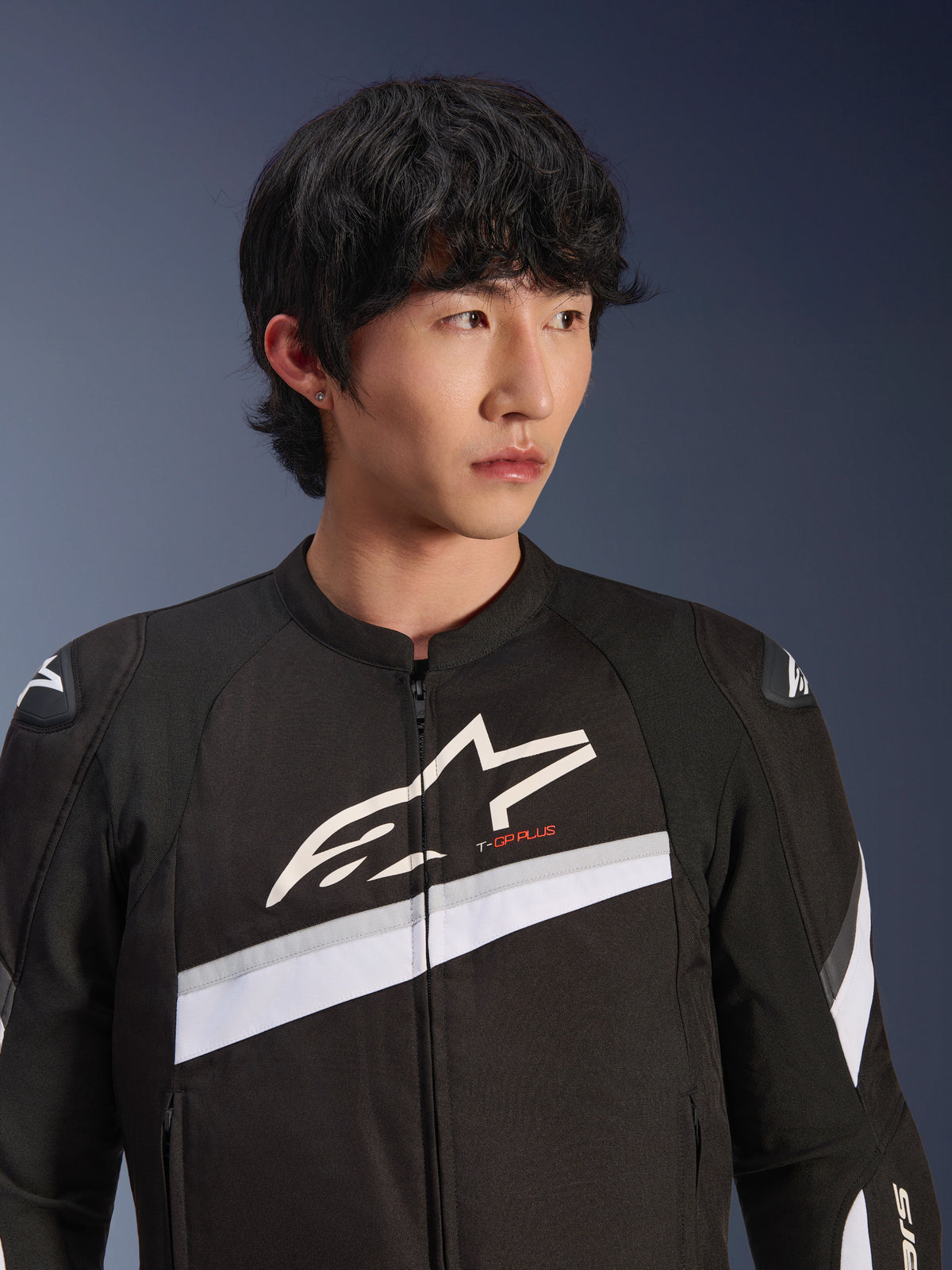 Kurtka Alpinestars T-GP Plus R V4, czarno-biała, tekstylna kurtka motocyklowa prezentowana na modelu, duże białe logo gwiazdy na klatce piersiowej, białe ukośne pasy, ochraniacze ramion DFS Lite, sportowa odzież motocyklowa do jazdy miejskiej.