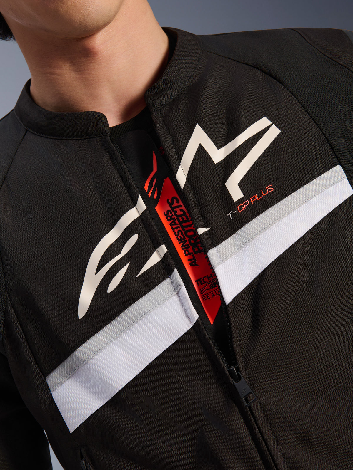 Kurtka Alpinestars T-GP Plus R V4, zbliżenie na klatkę piersiową i szyję, czarny materiał tekstylny z białym logo gwiazdy i oznaczeniem T-GP PLUS, biało-szary poziomy pas, rozpięty zamek odsłaniający czerwoną wewnętrzną patkę z napisem Tech-Air Ready