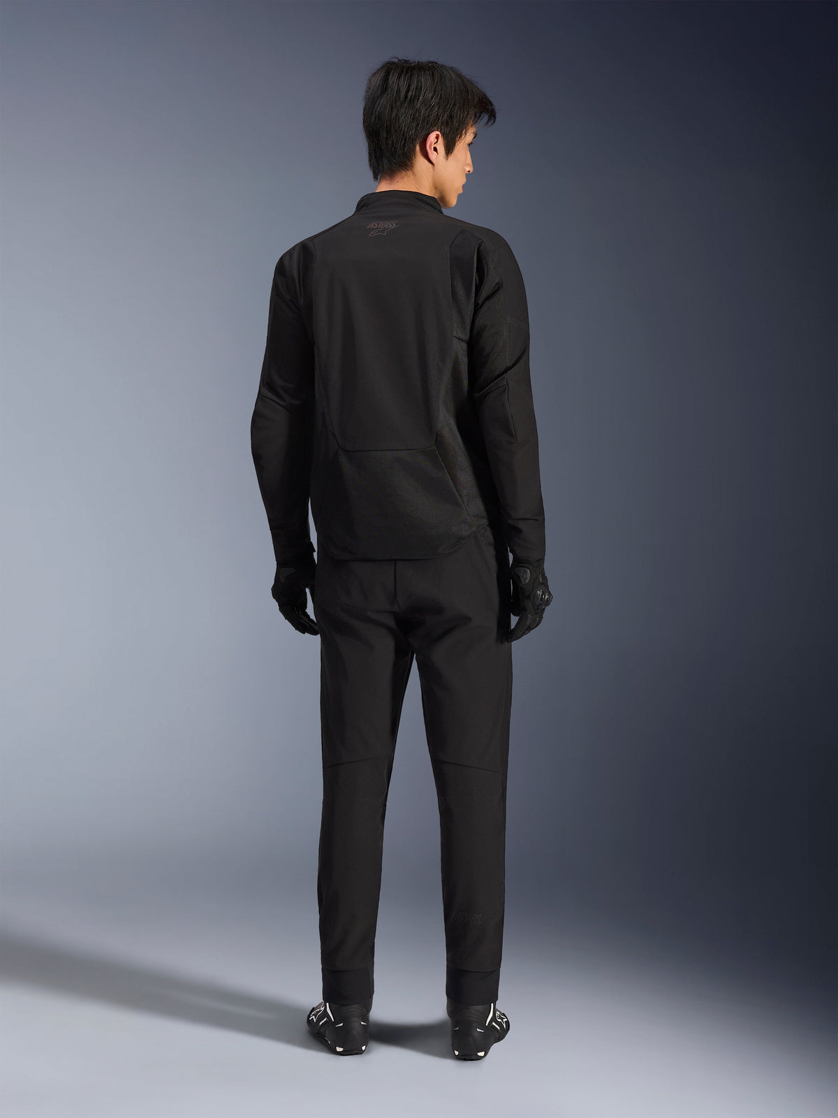 Alpinestars Aeroshell Airflow Jacket in Black Anthracite, back view of a male model wearing full motorcycle riding gear including black pants and gloves, featuring a lightweight softshell construction with an elongated rear for coverage.