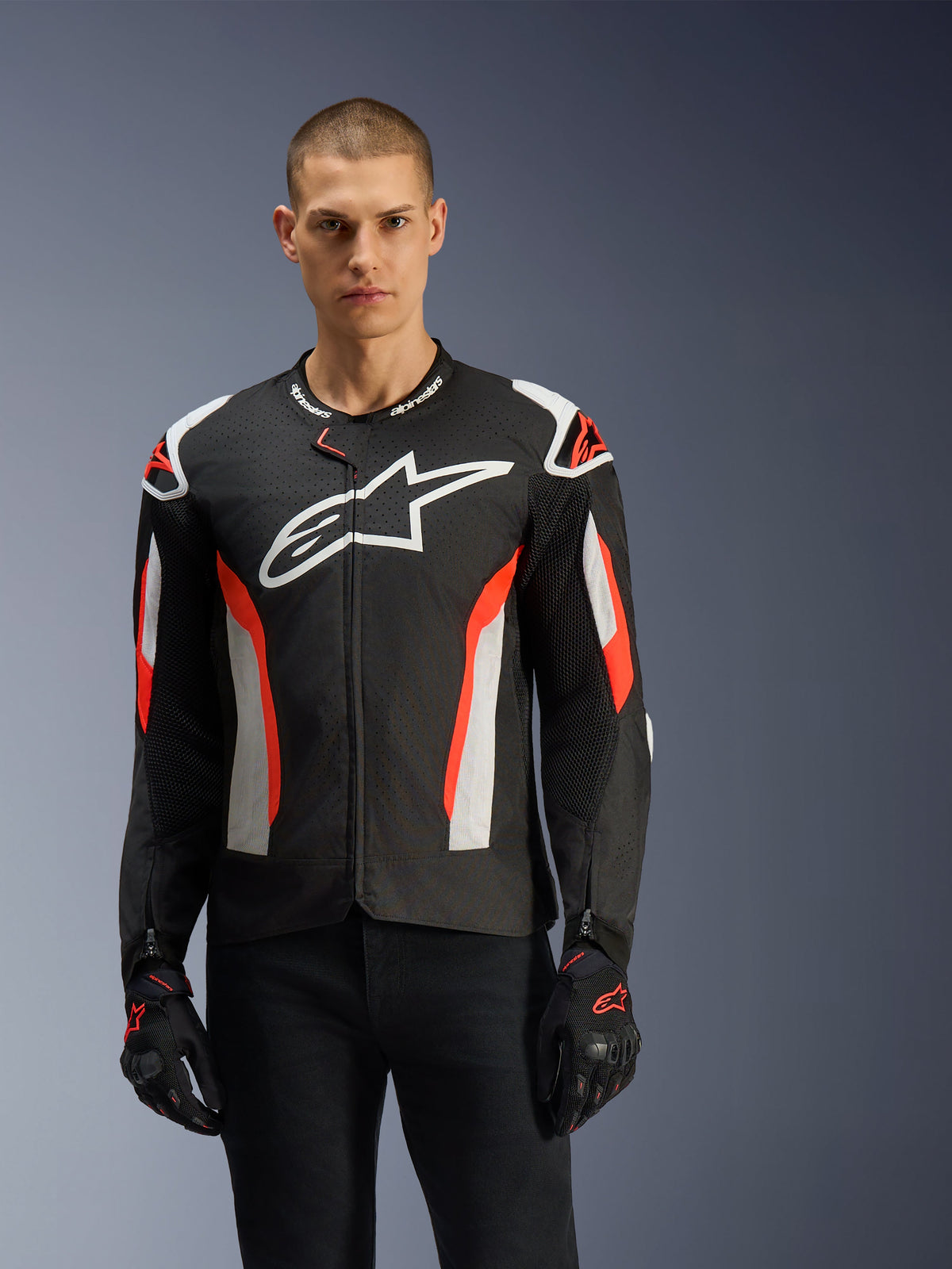 Male model wearing the Alpinestars T-GP Air Jacket, Black with Red Fluo and white accents, large Alpinestars logo on chest, featuring laser-perforated panels and shoulder armor, worn with black riding gloves, sport-cut motorcycle gear for warm weather