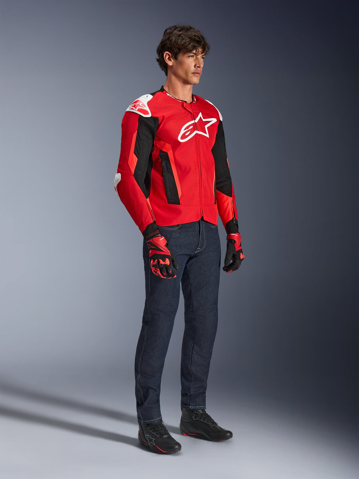 Alpinestars T-GP Air Jacket, Sport Motorcycle Jacket, Bright Red and Black with white accents, worn by a model with dark blue jeans and red racing gloves, featuring high-flow mesh panels and white shoulder sliders, aerodynamic sport fit for warm weather riding.