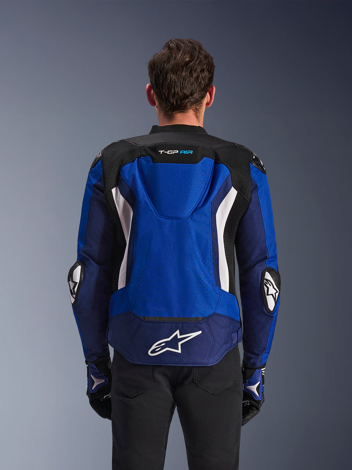 Alpinestars T-GP Air Jacket, sportowa tekstylna kurtka motocyklowa, jasnoniebieska, granatowa i biała, widok z tyłu prezentujący zintegrowany garb aerodynamiczny i duże białe logo w dolnej części pleców, noszona z dopasowanymi niebiesko-czarnymi rękawicami motocyklowymi