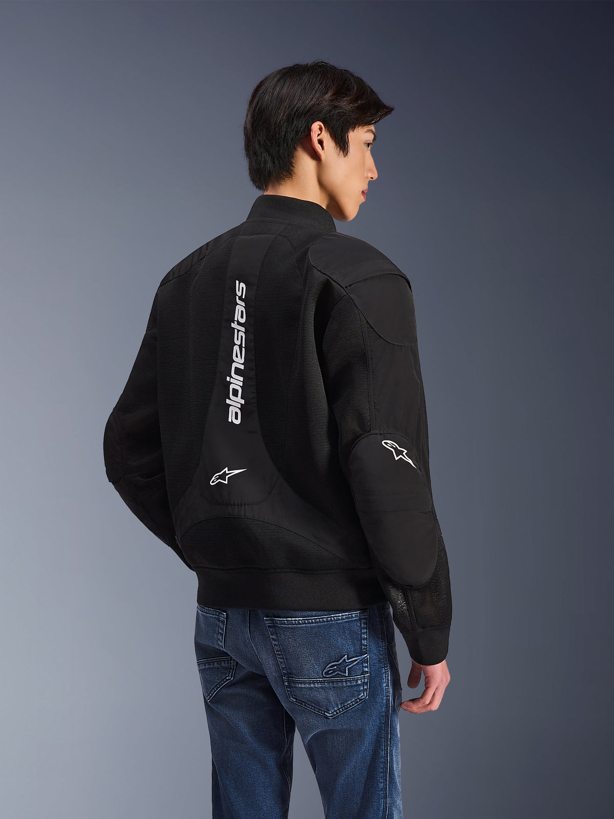 Kurtka Alpinestars Flight Air, czarna kurtka motocyklowa, widok z tyłu z dużym pionowym białym logo i siatkowymi panelami wentylacyjnymi, fason typu bomber z prążkowanym ściągaczem, zestawiona z niebieskimi dżinsami, nowoczesna miejska odzież motocyklowa
