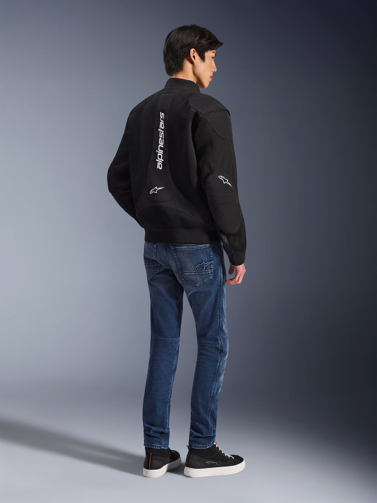 Model w czarnej kurtce Alpinestars Flight Air, widok z tyłu, w niebieskich jeansach motocyklowych i czarnych sneakersach, z pionowym białym logo Alpinestars i panelami wentylacyjnymi z siatki 3D.