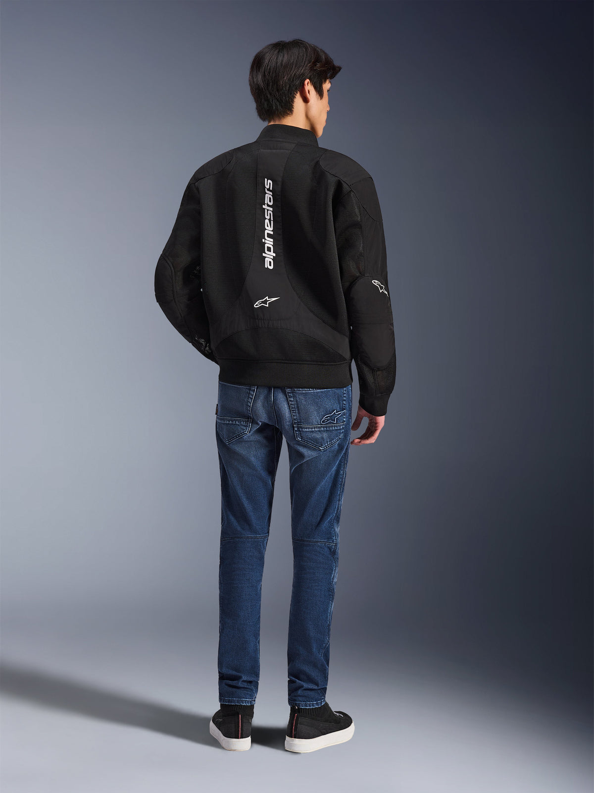 Model w kurtce Alpinestars Flight Air widoczny od tyłu, czarna kurtka motocyklowa w stylu bomber z panelami z siatki 3D i pionowym białym logo, niebieskie jeansowe spodnie motocyklowe i czarne sneakersy, współczesna miejska odzież motocyklowa