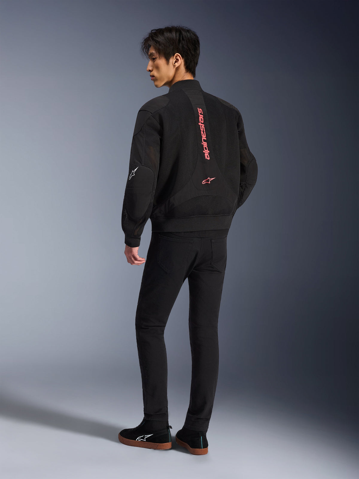 Męski model ubrany w kurtkę Alpinestars Flight Air w kolorze Black Bright Red, widok z tyłu ukazujący pionowe czerwone logo na plecach oraz konstrukcję z siatki 3D, w połączeniu z czarnymi spodniami i sneakersami, miejski styl motocyklowy
