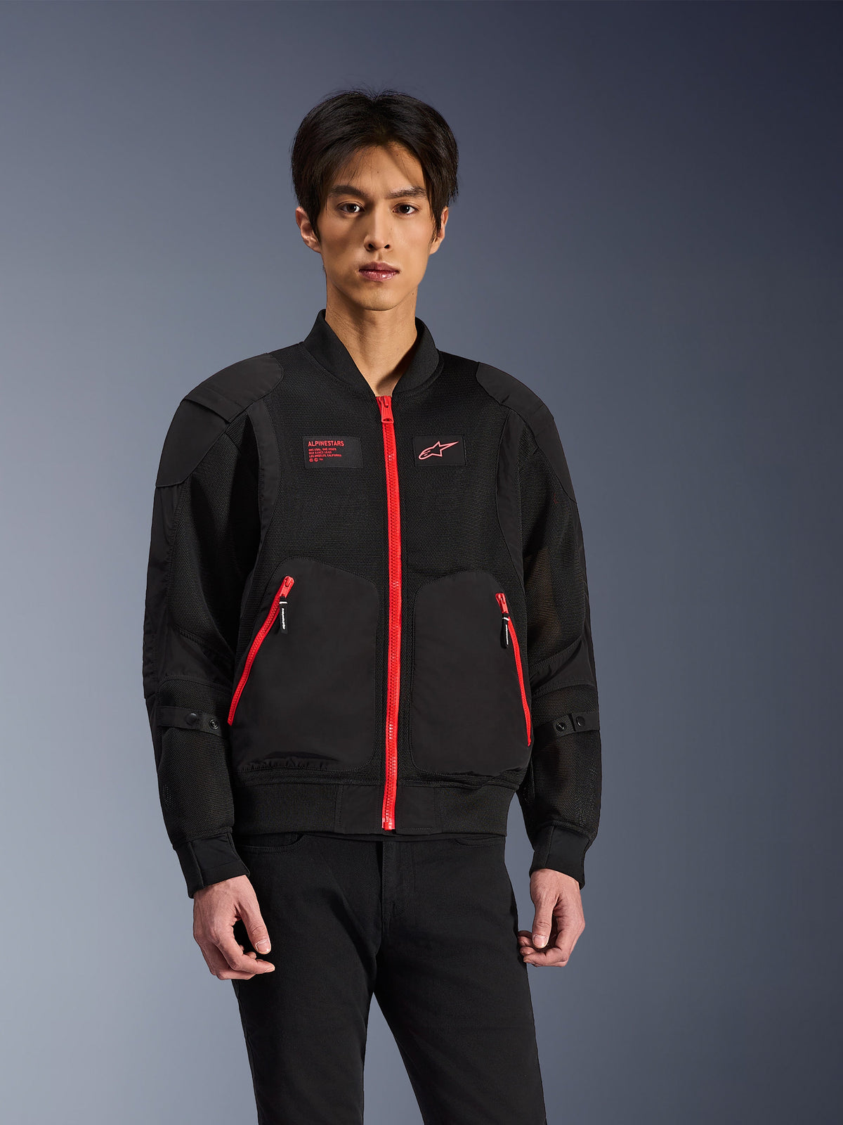 Alpinestars Flight Air Jacket w kolorze czarnym i jasnoczerwonym, miejska kurtka motocyklowa w stylu bomber noszona przez modela, z siatkowymi panelami wentylacyjnymi, wyrazistym czerwonym zamkiem błyskawicznym, czerwonymi detalami kieszeni i logo na piersi, zestawiona z czarnymi spodniami