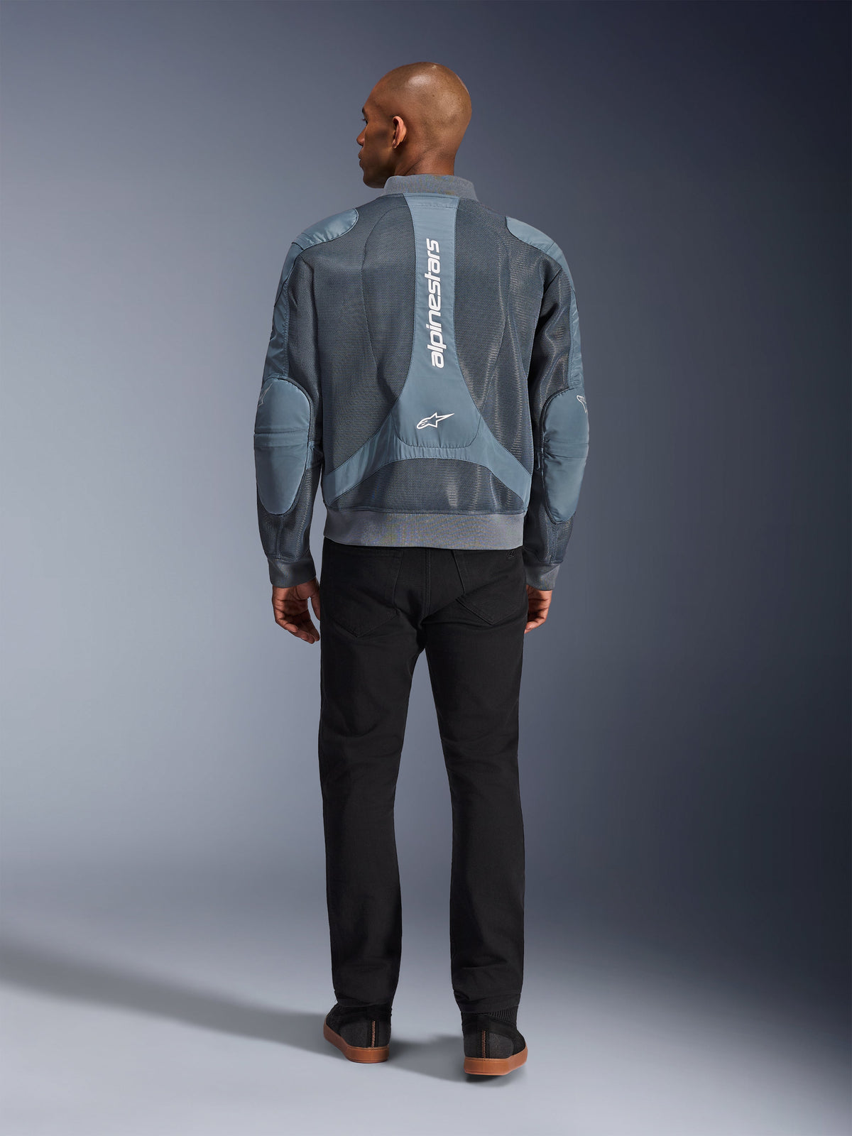 Kurtka Alpinestars Flight Air w kolorze Storm Gray, widok z tyłu na modela w miejskiej kurtce motocyklowej z panelami z siatki 3D i pionowym logo Alpinestars, w zestawieniu z czarnymi spodniami tworzącymi nowoczesny, miejski styl.