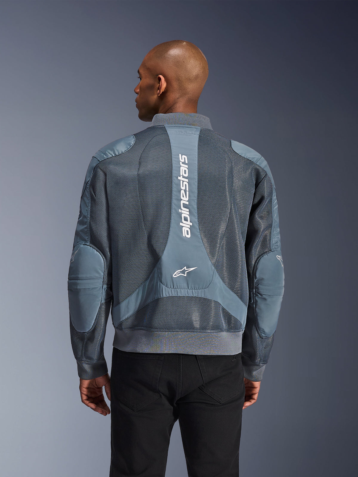 Alpinestars Flight Air Jacket w kolorze Storm Gray, widok z tyłu na modelu, miejska kurtka motocyklowa z rozszerzonymi panelami z siatki 3D, pionowym logo Alpinestars na plecach oraz ściągaczami u dołu i przy kołnierzu w stylu bomberki.