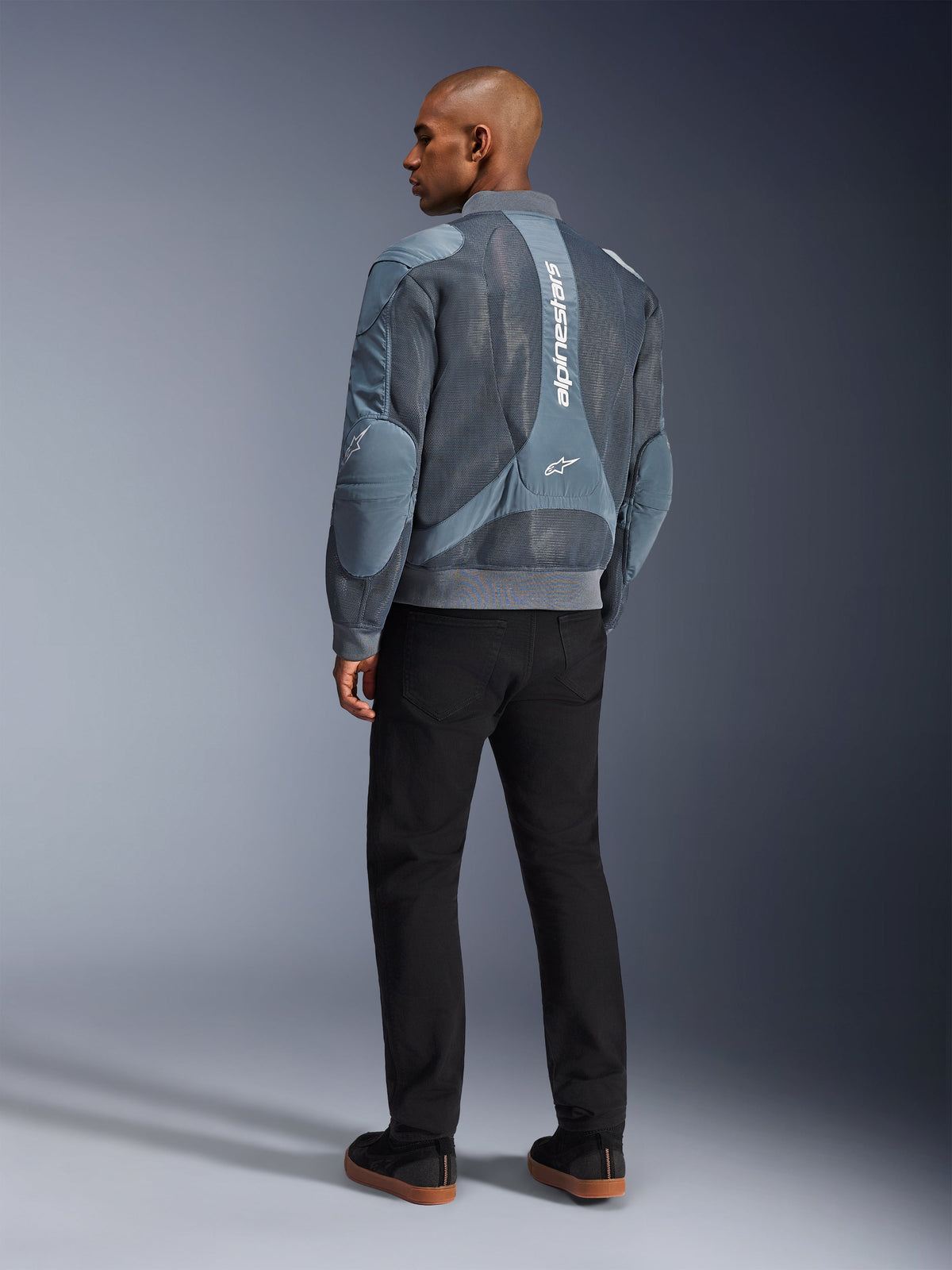 Alpinestars Flight Air Jacket, kurtka motocyklowa Storm Gray, widok z tyłu, z panelami wentylacyjnymi z siatki 3D i pionowym białym logo Alpinestars, noszona z czarnymi spodniami i czarnymi sneakersami, miejski styl commuter