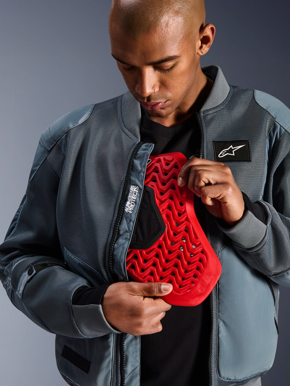 Alpinestars Flight Air Jacket w kolorze Storm Gray, miejska motocyklowa kurtka typu bomber z panelami z siatki 3D zapewniającymi przepływ powietrza, rozpięta, by odsłonić czerwoną wkładkę ochraniacza pleców Nucleon, z brandingiem „Alpinestars Protects” i nowoczesną estetyką streetwear