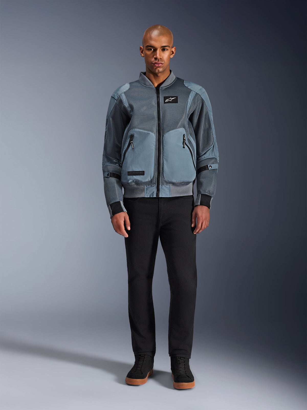 Alpinestars Flight Air Jacket w kolorze Storm Gray, miejska kurtka motocyklowa inspirowana stylem bomber, panele z siatki 3D i jednolite przednie kieszenie, noszona przez mężczyznę w czarnych spodniach i czarnych wysokich butach, nowoczesna odzież motocyklowa street-style