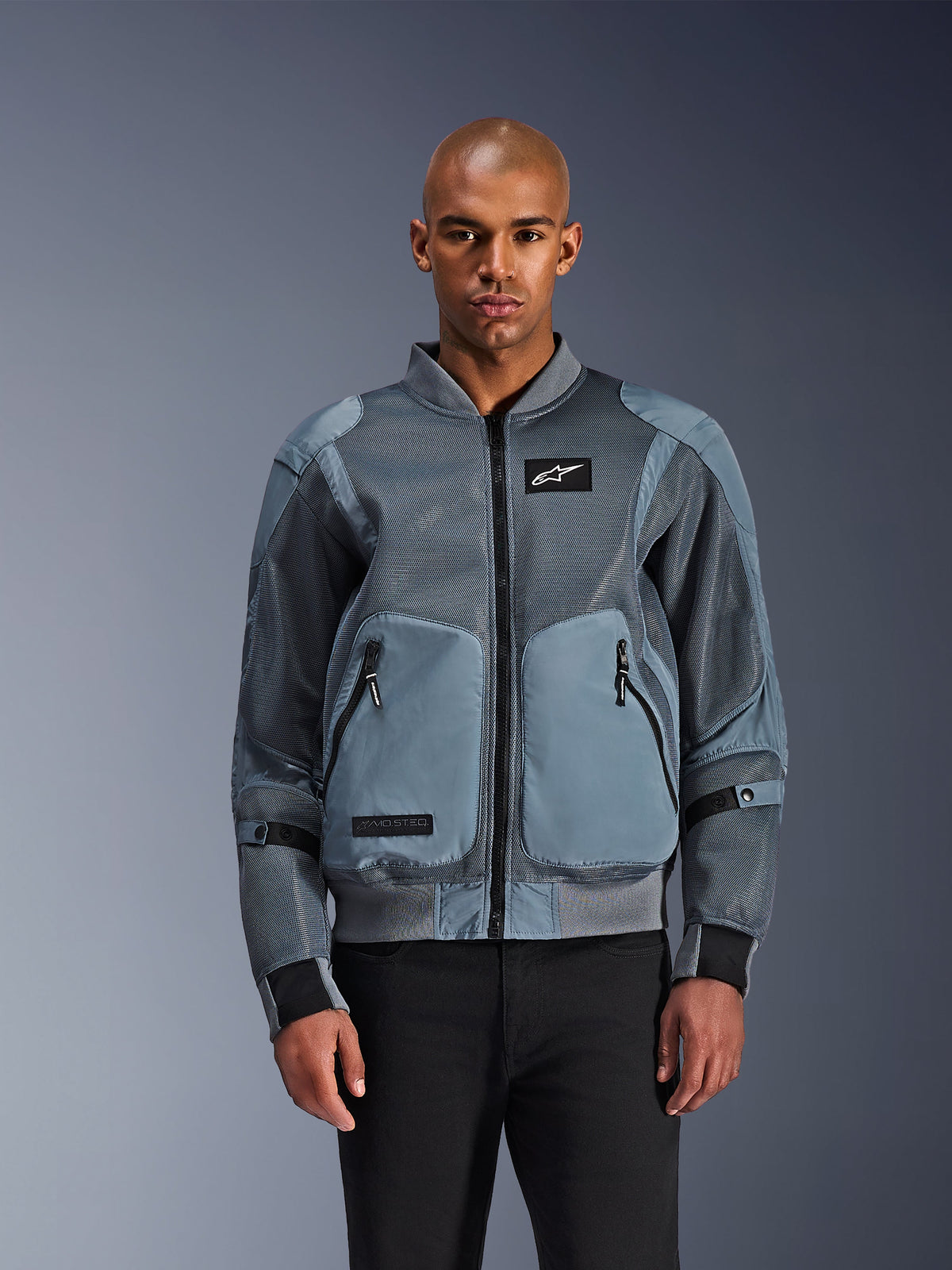 Alpinestars Flight Air Jacket, miejska kurtka motocyklowa typu bomber, kolor Storm Gray, posiada panele wentylacyjne z siatki 3D i białą naszywkę z logo Astars, noszona przez modela z czarnymi spodniami, nowoczesna miejska odzież motocyklowa