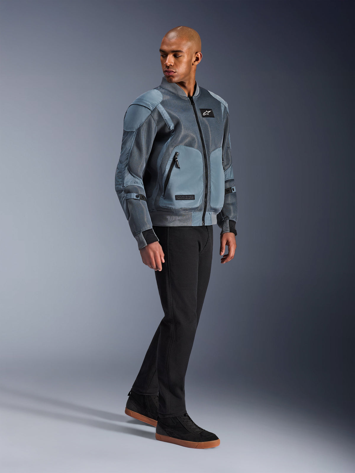 Kurtka Alpinestars Flight Air, Storm Gray, miejska kurtka motocyklowa inspirowana stylem bomber, noszona przez modela z czarnymi spodniami i sneakersami, wyposażona w rozszerzone panele wentylacyjne z siatki 3D oraz zewnętrzne kieszenie na ochraniacze, nowoczesna odzież ochronna streetwear do jazdy w upalne dni
