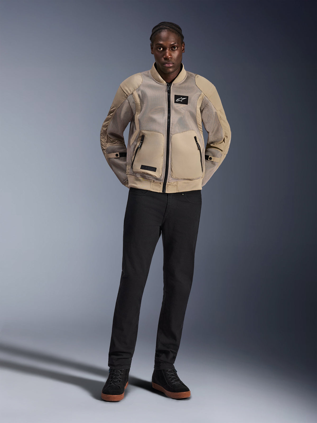 Model w kurtce Alpinestars Flight Air w kolorze brązowym, miejska kurtka motocyklowa z rozbudowanymi panelami z siatki 3D, kołnierz i mankiety ze ściągaczami w stylu bomber, zestawiona z czarnymi spodniami i czarnymi butami motocyklowymi, współczesna odzież w stylu ulicznym