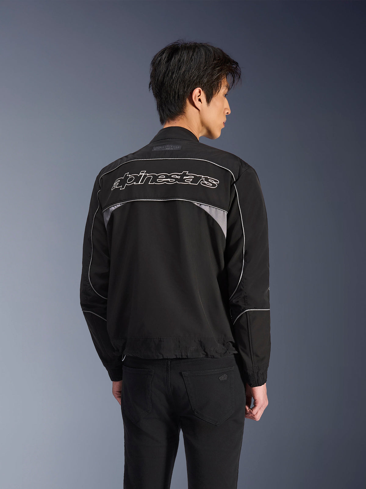 Alpinestars Unite Jacket, miejska kurtka motocyklowa, czarna z ciemnoszarymi akcentami, widok z tyłu pokazujący biały napis Alpinestars na ramionach, odblaskowe lamówki i panele, minimalistyczny design softshell streetwear
