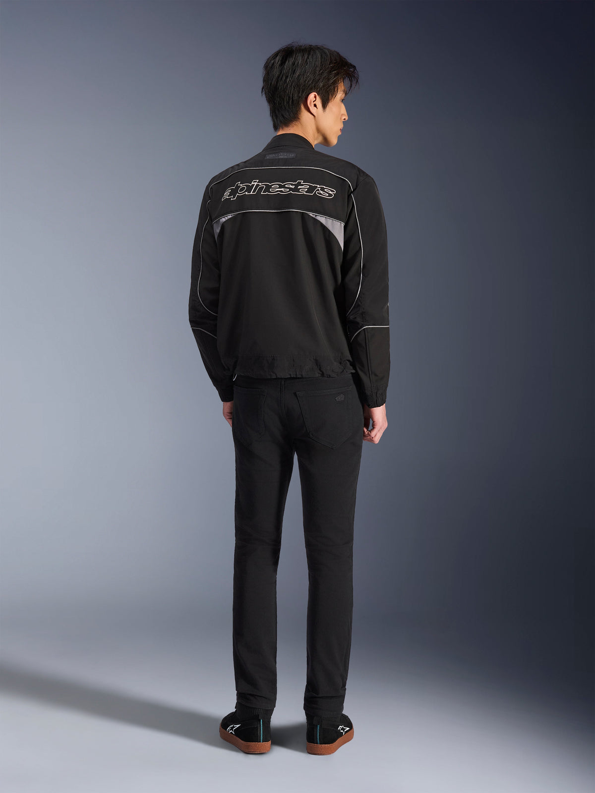 Kurtka Alpinestars Unite, czarno-ciemnoszara kurtka motocyklowa, widok z tyłu prezentujący konturowe logo Alpinestars na plecach, szare panele odblaskowe, minimalistyczny miejski design, noszona z czarnymi spodniami i czarnymi butami.