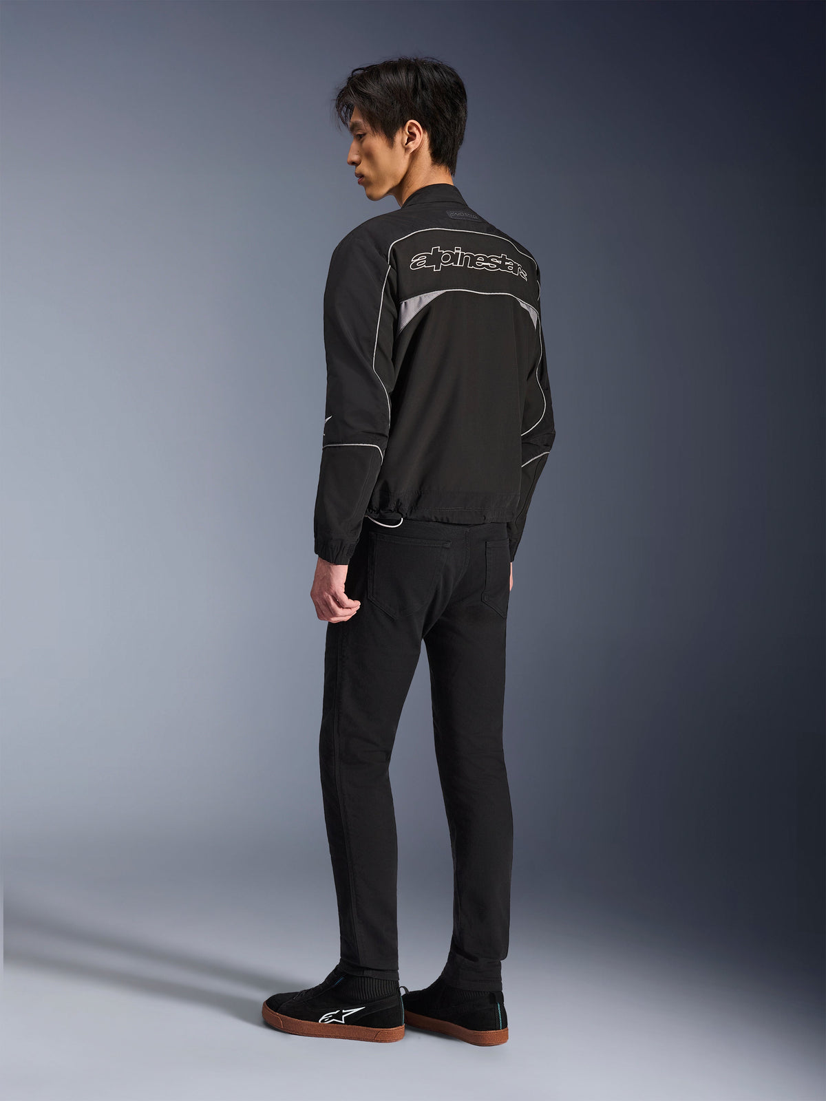 Kurtka Alpinestars Unite, miejska kurtka motocyklowa, czarna i ciemnoszara, widok z tyłu przedstawiający duże konturowe logo Alpinestars, odblaskowe lamówki i panele, minimalistyczny design softshell, noszona z czarnymi spodniami i codziennym obuwiem