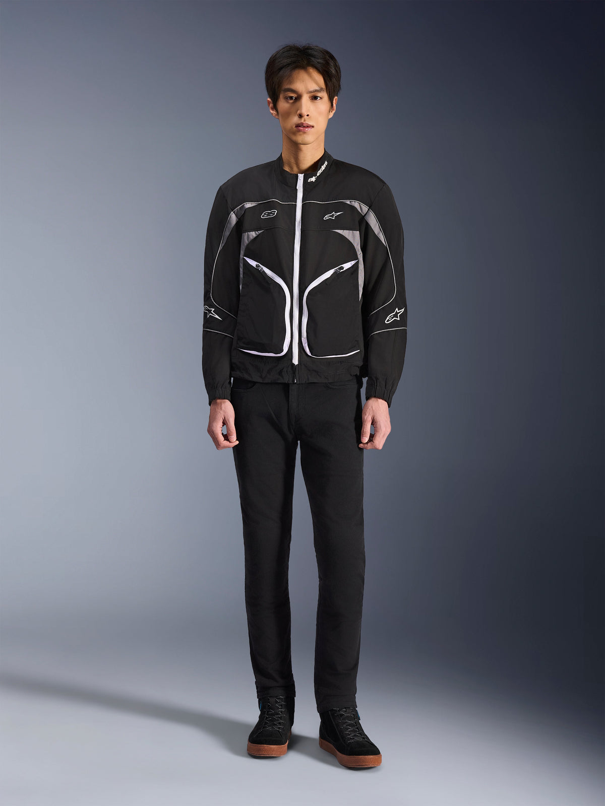 Kurtka Alpinestars Unite, kurtka motocyklowa, czarno-ciemnoszara, prezentowana przez modela w czarnych spodniach i czarnych butach, z białymi kontrastowymi zamkami, odblaskowymi lamówkami i minimalistycznym miejskim designem