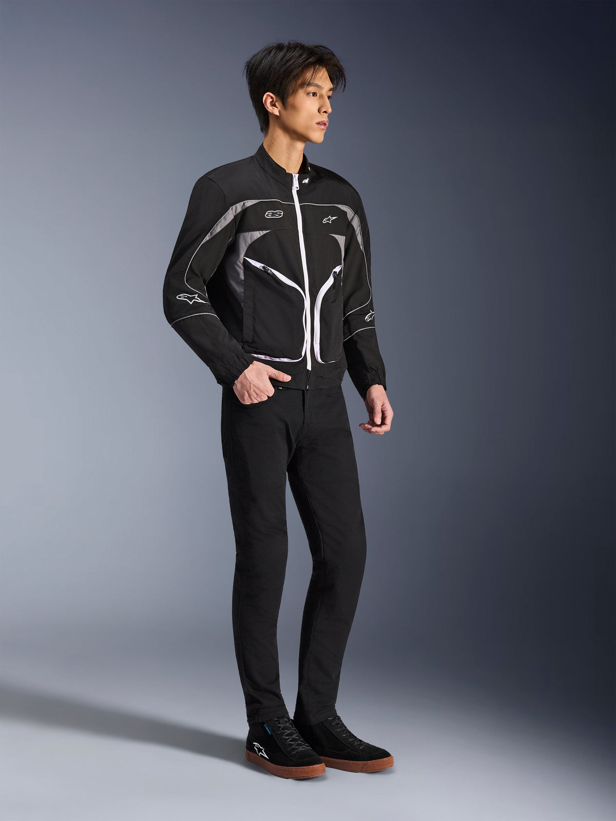 Model w kurtce Alpinestars Unite Jacket w kolorze czarnym i ciemnoszarym, miejska kurtka motocyklowa z białymi zamkami i odblaskowymi detalami, w zestawieniu z czarnymi spodniami slim-fit i sneakersami, minimalistyczna odzież motocyklowa inspirowana stylem ulicznym