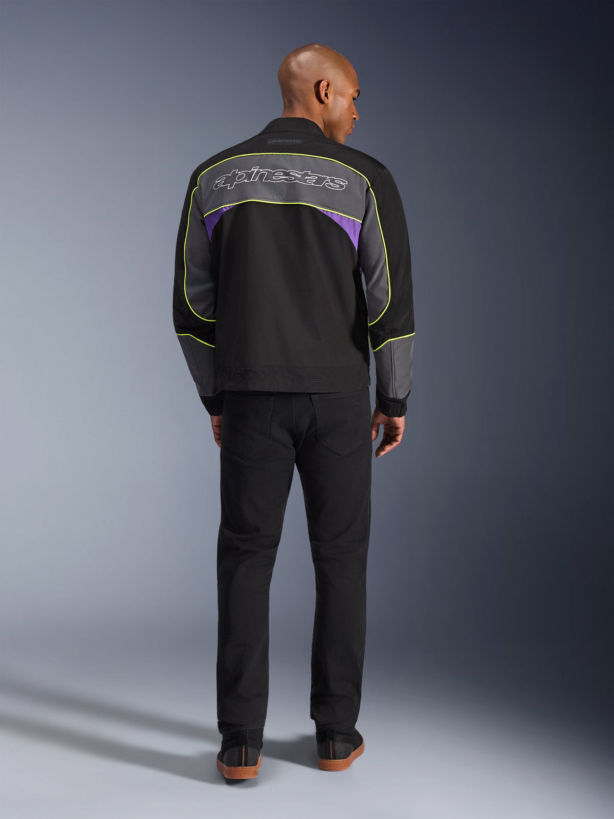 Kurtka Alpinestars Unite, miejska kurtka motocyklowa w kolorze Black Asphalt Violet, widok z tyłu prezentujący szare panele z fioletowymi akcentami i neonowożółtymi lamówkami, logotyp Alpinestars na ramionach, noszona z czarnymi spodniami