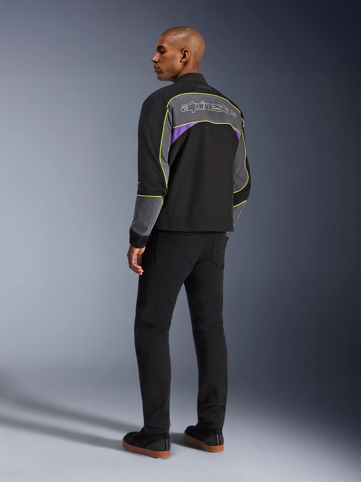 Kurtka Alpinestars Unite, miejska kurtka motocyklowa, kolor Black Asphalt Violet, widok z tyłu pod kątem na modelu, czarny softshell z szarymi panelami i fioletowymi akcentami, neonowożółte odblaskowe lamówki, logo Alpinestars w górnej części pleców, w zestawieniu z czarnymi spodniami i czarnymi butami typu casual