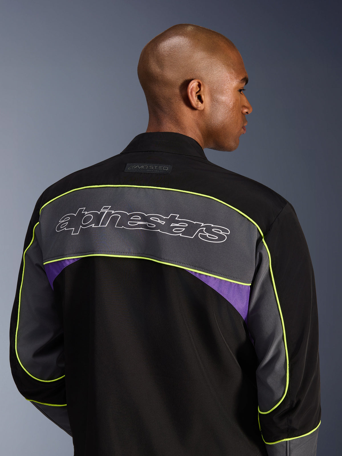 Kurtka Alpinestars Unite, miejska kurtka motocyklowa, Black Asphalt Violet, widok z tyłu pokazujący duży szary panel z logo Alpinestars, neonowożółte lamówki i fioletowe akcenty na 3-warstwowej konstrukcji softshell