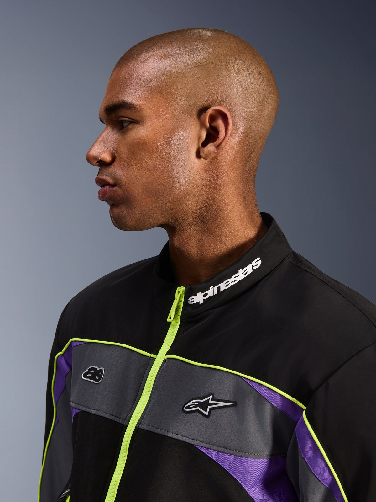 Alpinestars Unite Jacket, Black Asphalt Violet, widok z profilu na modelu męskim, skupienie na wysokim kołnierzu z białym logo Alpinestars, neonowożółty zamek, szare panele na klatce piersiowej z logo, fioletowe akcenty, minimalistyczna miejska stylistyka motocyklowa streetwear