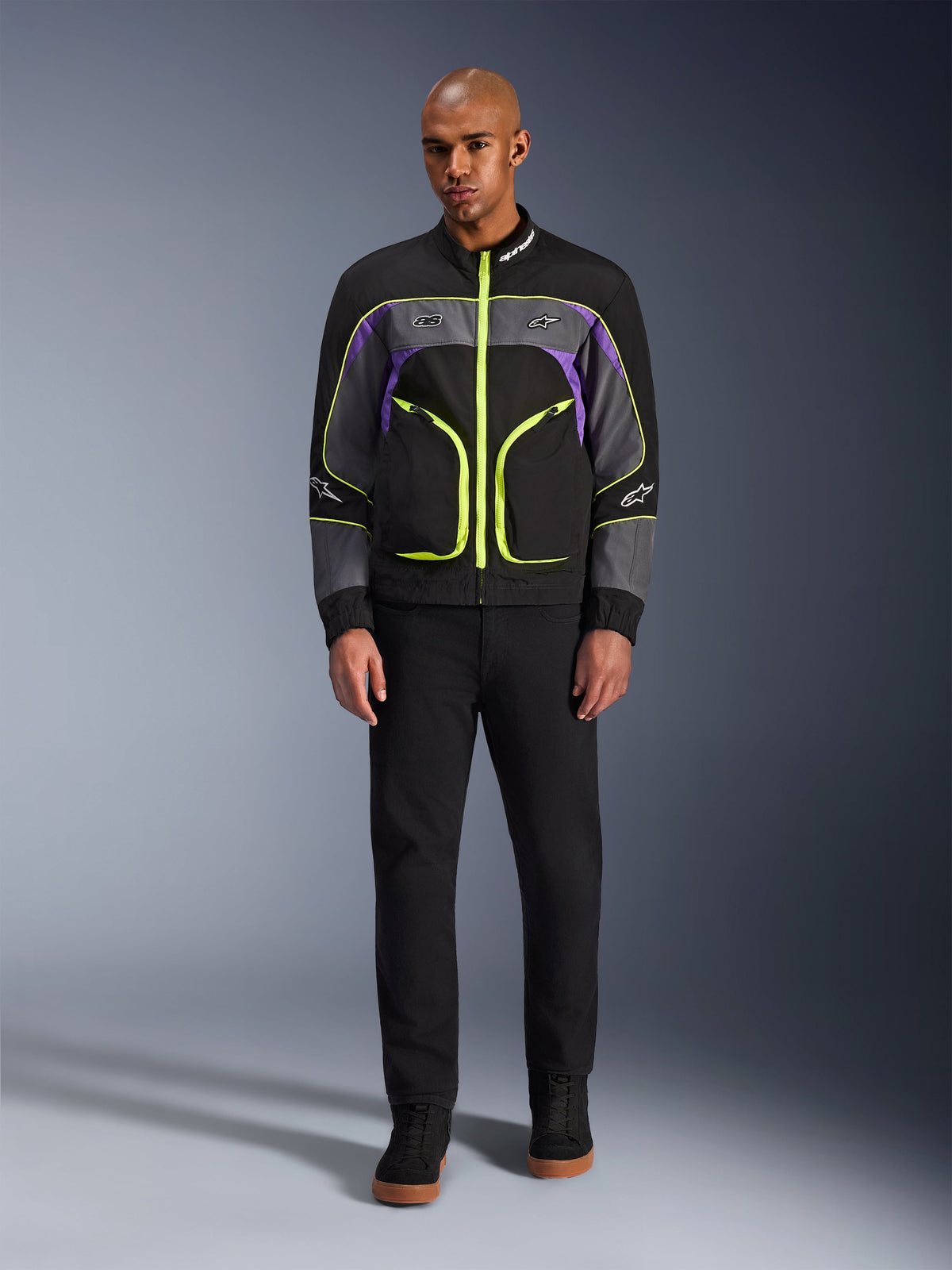 Męski model w kurtce Alpinestars Unite w kolorze Black Asphalt Violet, czarna baza z szarymi i fioletowymi panelami, jaskrawe neonowo-żółte zamki i lamówki, w zestawieniu z czarnymi spodniami i butami, minimalistyczna miejska kurtka motocyklowa