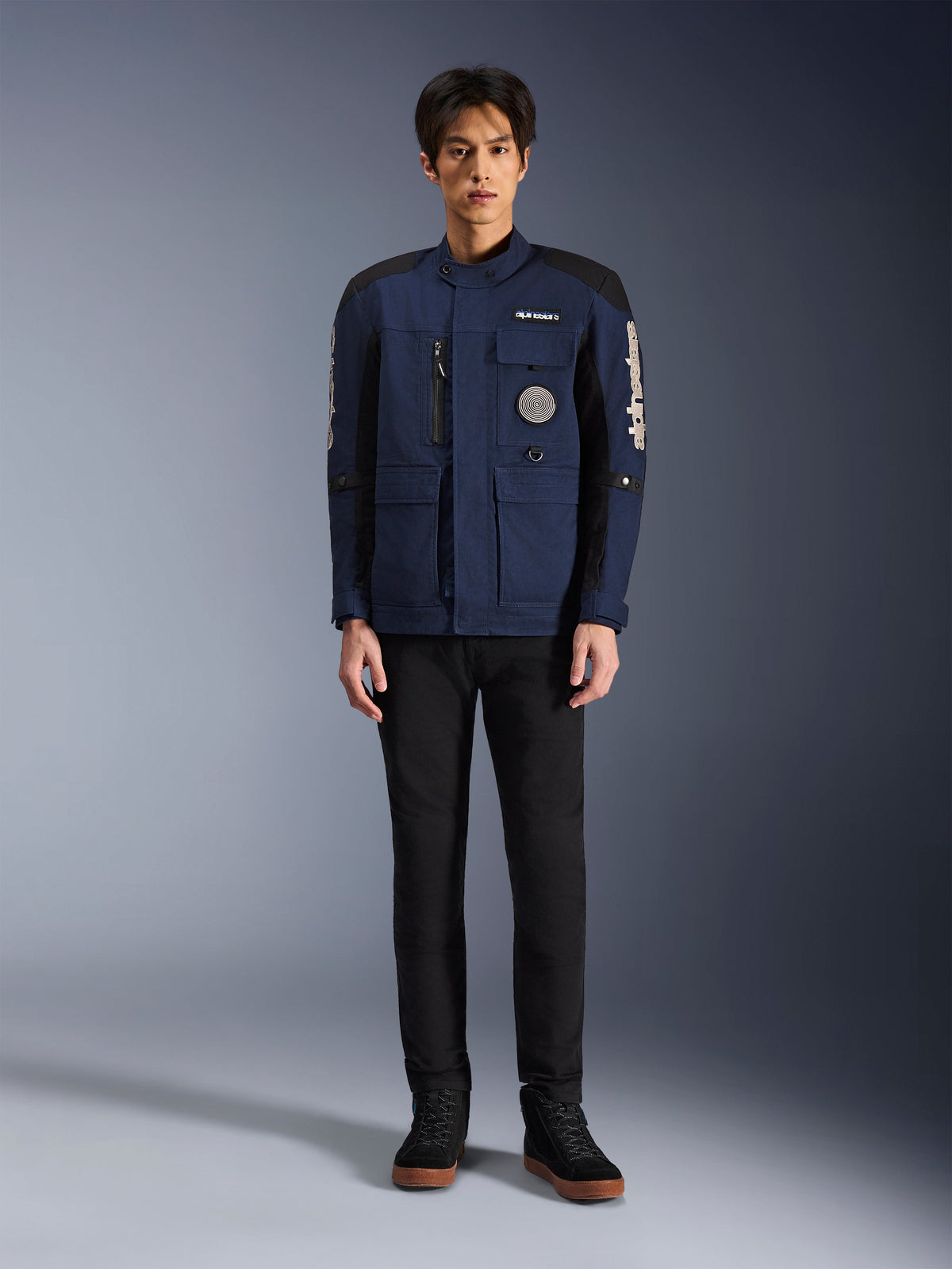 Alpinestars Dice Jacket, Urban Motorcycle Jacket, Dark Blue and Black, worn by a model with black pants and black shoes, modern field jacket design with multiple pockets, vertical chest zipper, and white Alpinestars branding on the sleeves.