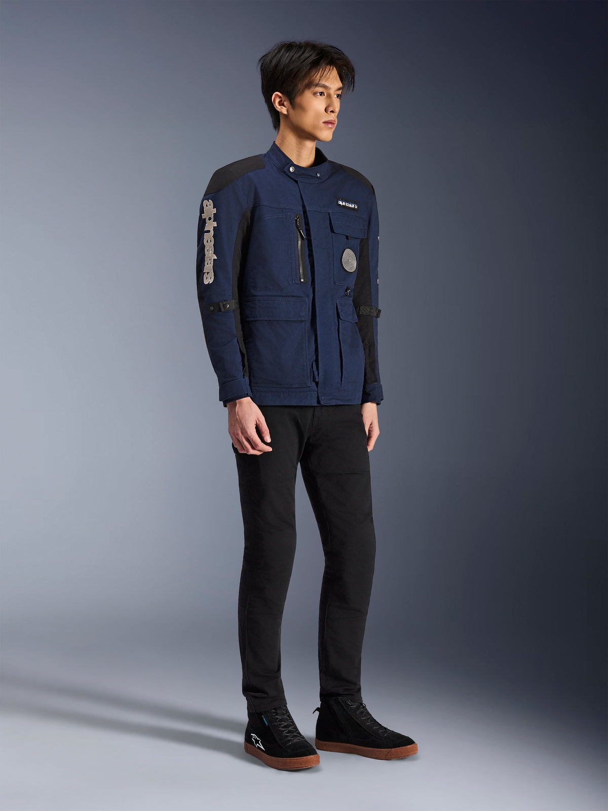 Alpinestars Dice Jacket, Dark Blue and Black, modern field-style motorcycle jacket worn by a model with black pants and black high-top sneakers, featuring multiple utility pockets, vertical Alpinestars branding on the sleeve, and woven patches, urban contemporary riding gear