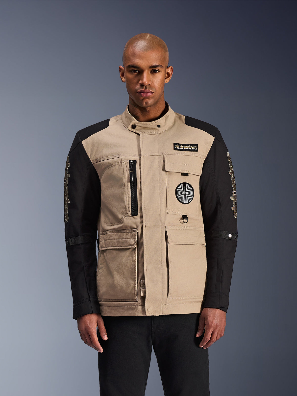 Alpinestars Dice Jacket, miejska kurtka motocyklowa, beżowo-czarna, surowa estetyka odzieży roboczej z licznymi kieszeniami użytkowymi i naszywkami na piersi, wysoki kołnierz zapinany na guzik, wyraziste logo na rękawach, prezentowana przez modela