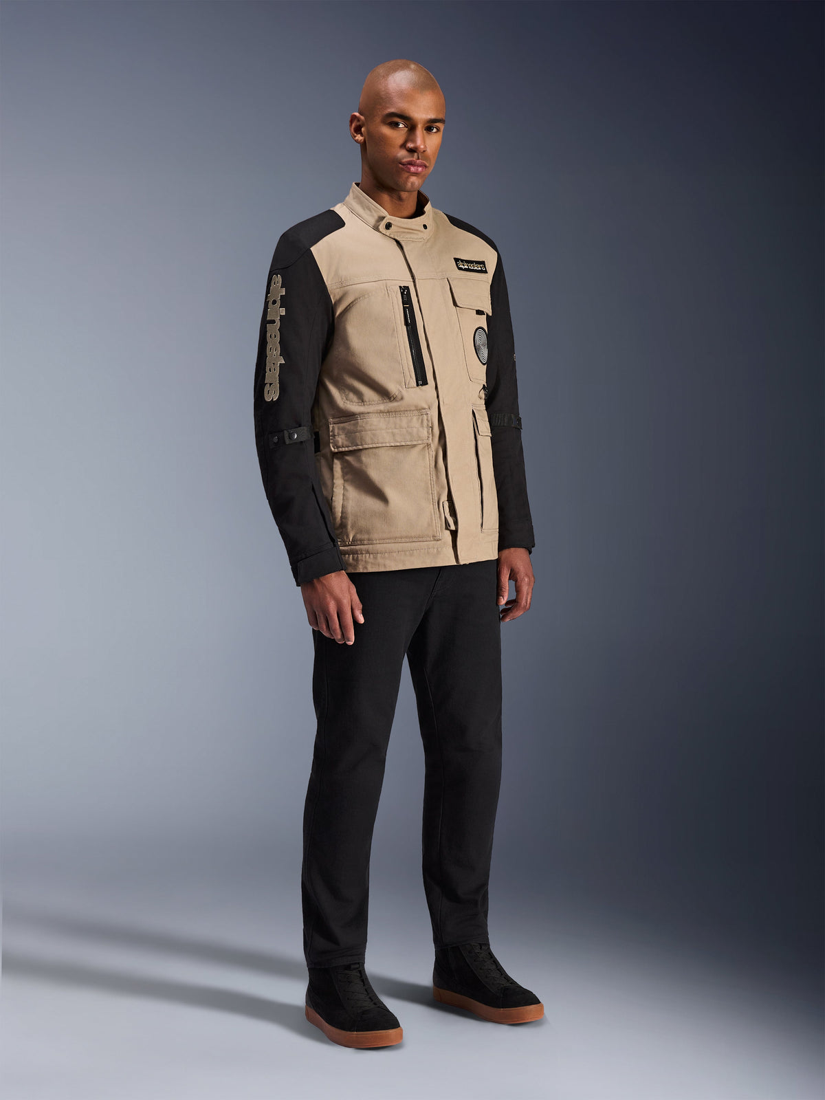 Alpinestars Dice Jacket, miejska kurtka motocyklowa, czarno-piaskowa, dwukolorowy design z czarnymi rękawami i korpusem w kolorze khaki, noszona przez modela w czarnych spodniach i czarnych butach sportowych, estetyka inspirowana odzieżą roboczą z kieszeniami użytkowymi i haftowanym logo na rękawie.