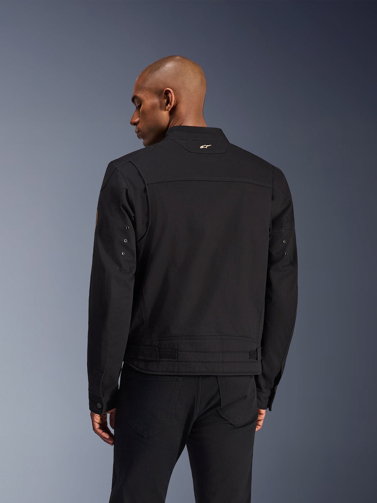 Kurtka Alpinestars Bruiser, czarna miejska kurtka motocyklowa, widok z tyłu, design inspirowany odzieżą roboczą, małe złote logo na kołnierzu i regulowane paski w talii, wytrzymała konstrukcja tekstylna do jazdy miejskiej