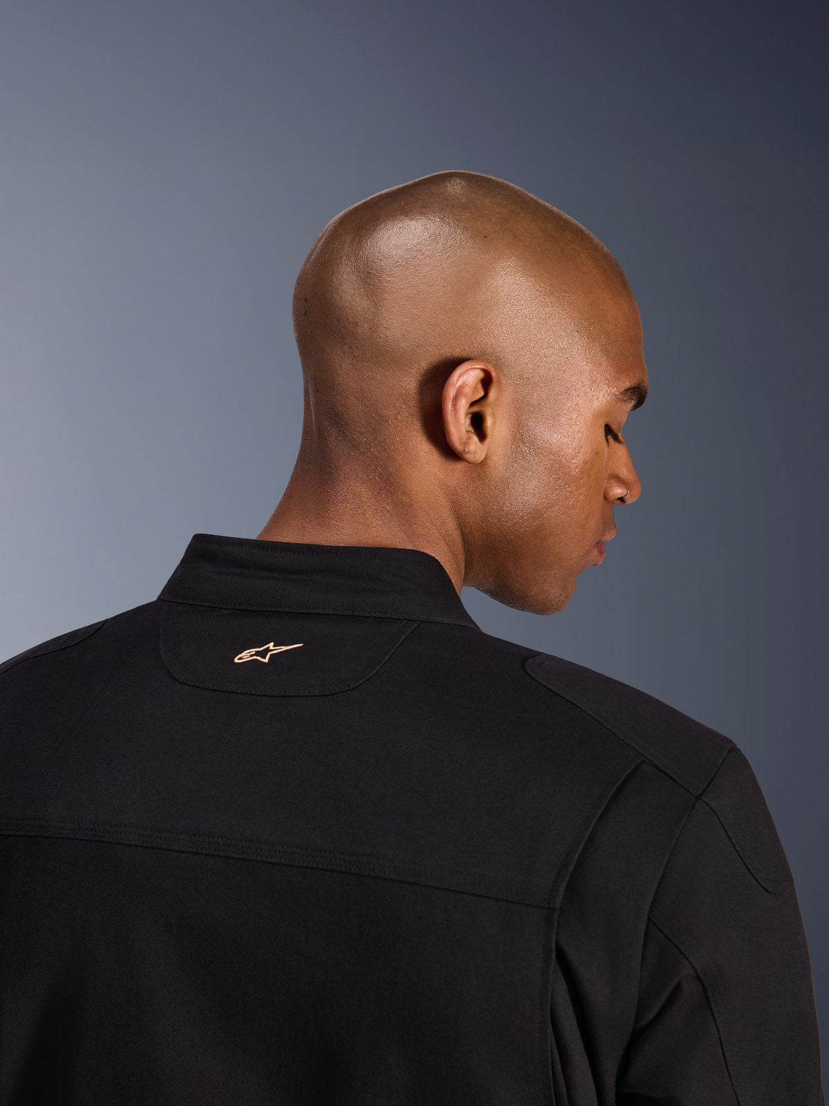 Alpinestars Bruiser Jacket, czarna, zbliżenie na tył kołnierza i górną część ramion, małe haftowane jasnobrązowe logo Alpinestars na karku, miejska kurtka motocyklowa w estetyce odzieży roboczej