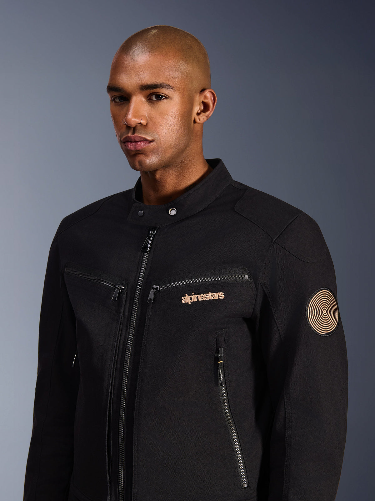 Męski model w kurtce Alpinestars Bruiser, czarna miejska kurtka motocyklowa z polibawełnianego twillu, wyposażona w kieszenie na zamek, złote haftowane logo na piersi oraz naszywkę Oscar spiral na rękawie, nowoczesny styl miejski