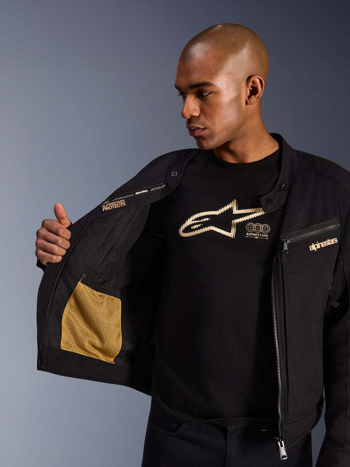 Alpinestars Bruiser Jacket, czarna miejska kurtka motocyklowa, zbliżenie na otwarte wnętrze ze złotą kieszenią z siateczki i napisem „Alpinestars Protects”, założona na czarny t-shirt z logo, tekstylna odzież motocyklowa o estetyce odzieży roboczej