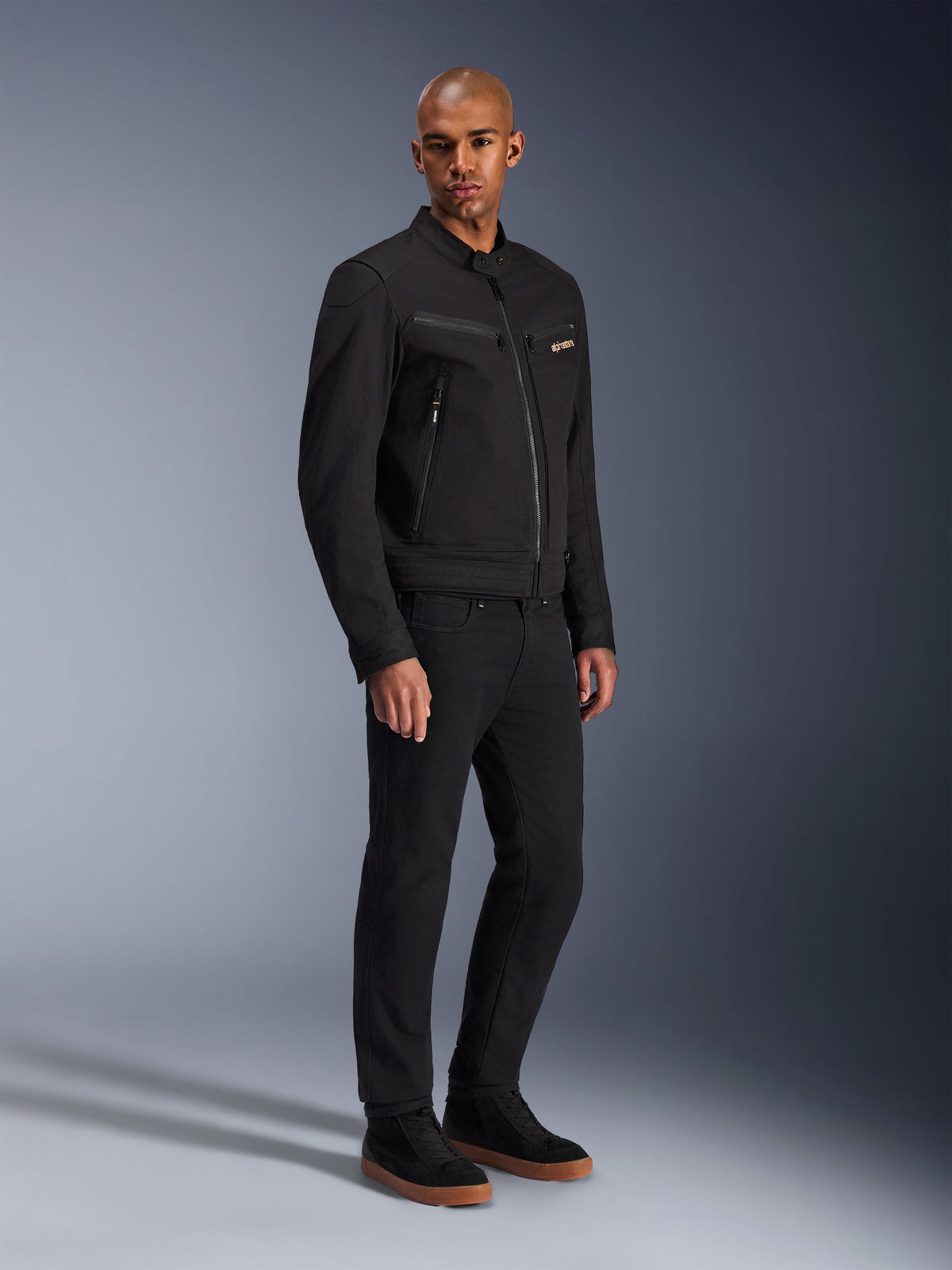 Alpinestars Bruiser Jacket, miejska kurtka motocyklowa, czarna, noszona przez modela w czarnych spodniach i butach sportowych, z kieszeniami na piersi zapinanymi na zamek, tkanina polycotton twill, elegancki design inspirowany odzieżą roboczą do jazdy miejskiej