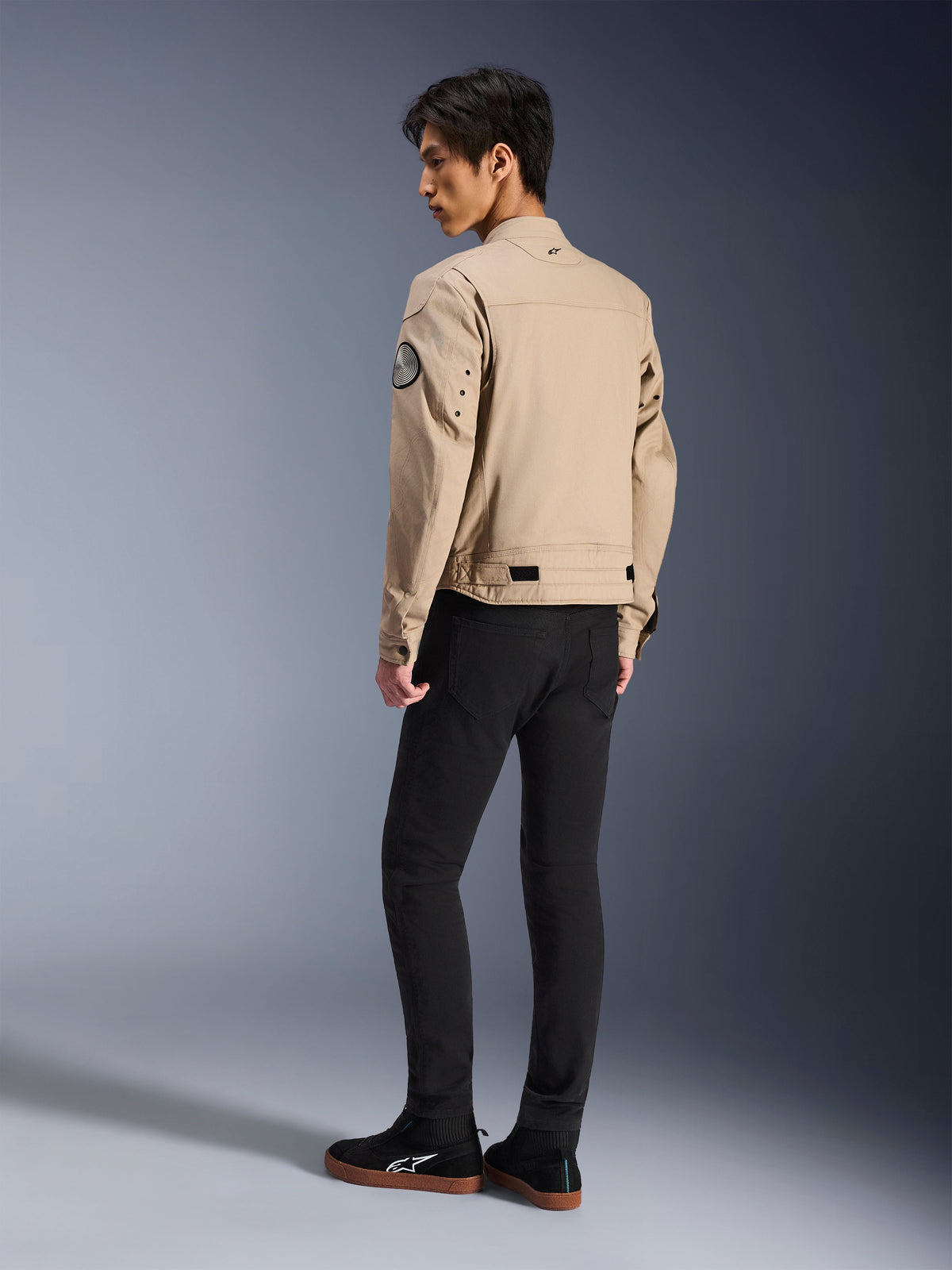 Alpinestars Bruiser Jacket, męski model widoczny od tyłu, beżowa miejska kurtka motocyklowa w stylistyce odzieży roboczej, okrągła naszywka na rękawie, paski do regulacji w talii i profilowane ramiona, czarne spodnie i sneakersy