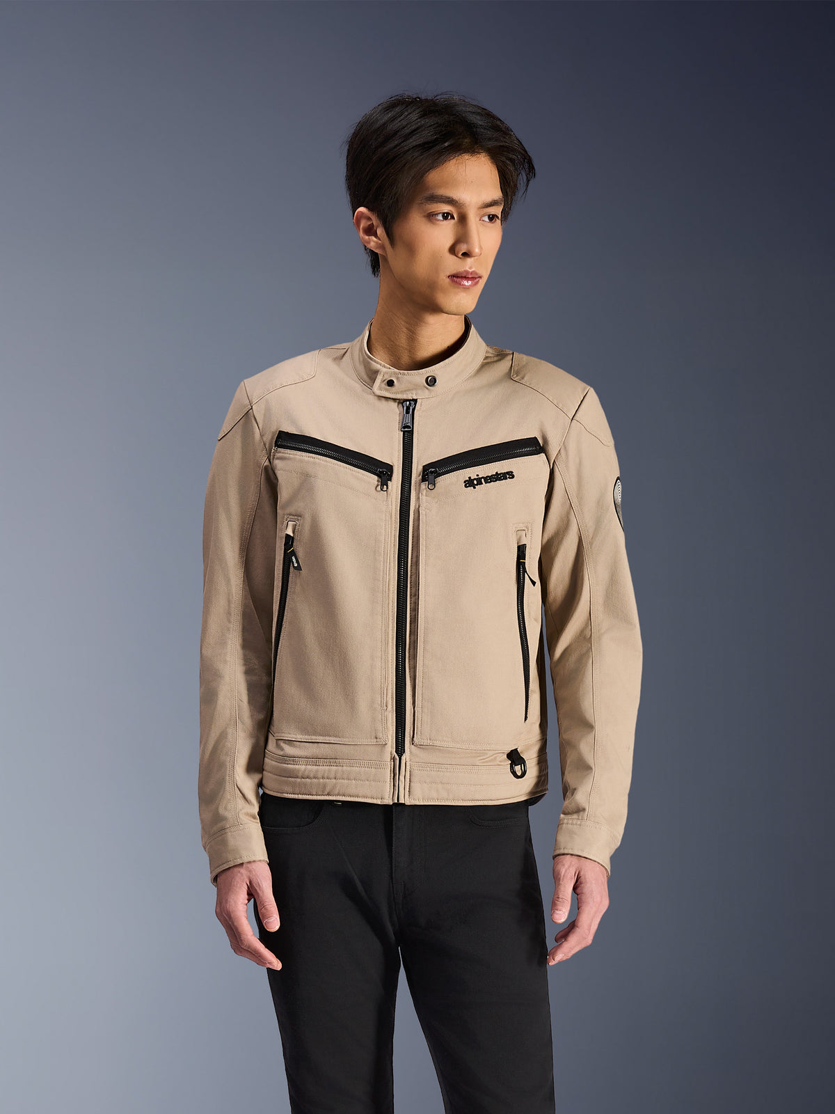 Alpinestars Bruiser Jacket, beżowa kurtka motocyklowa noszona przez modela, czarne detale zamków na klatce piersiowej i bocznych kieszeniach, wysoki kołnierz na zatrzaski, miejska odzież motocyklowa inspirowana stylem roboczym, w zestawieniu z czarnymi spodniami